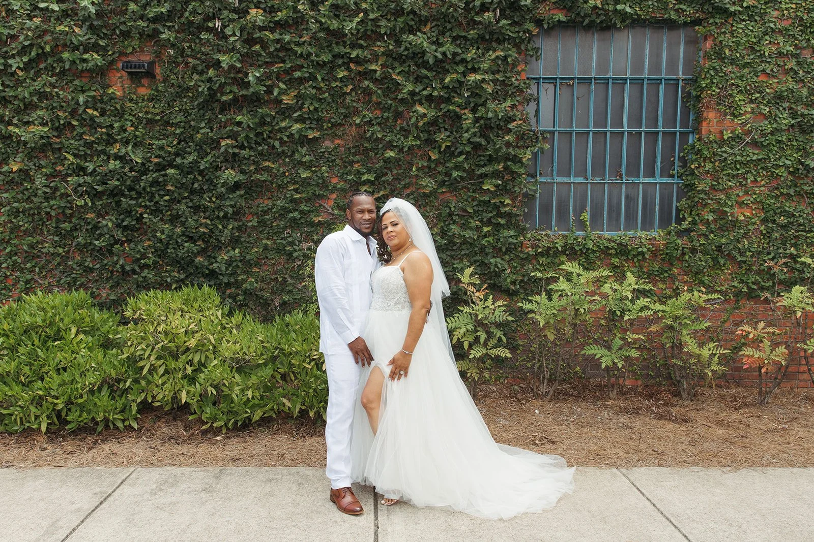 Bride and groom posing in front of the signature green ivy wall at Studio 215 in downtown Fayetteville, NC; a lush urban garden photography backdrop at our industrial warehouse venue near Fort Bragg and Raleigh.