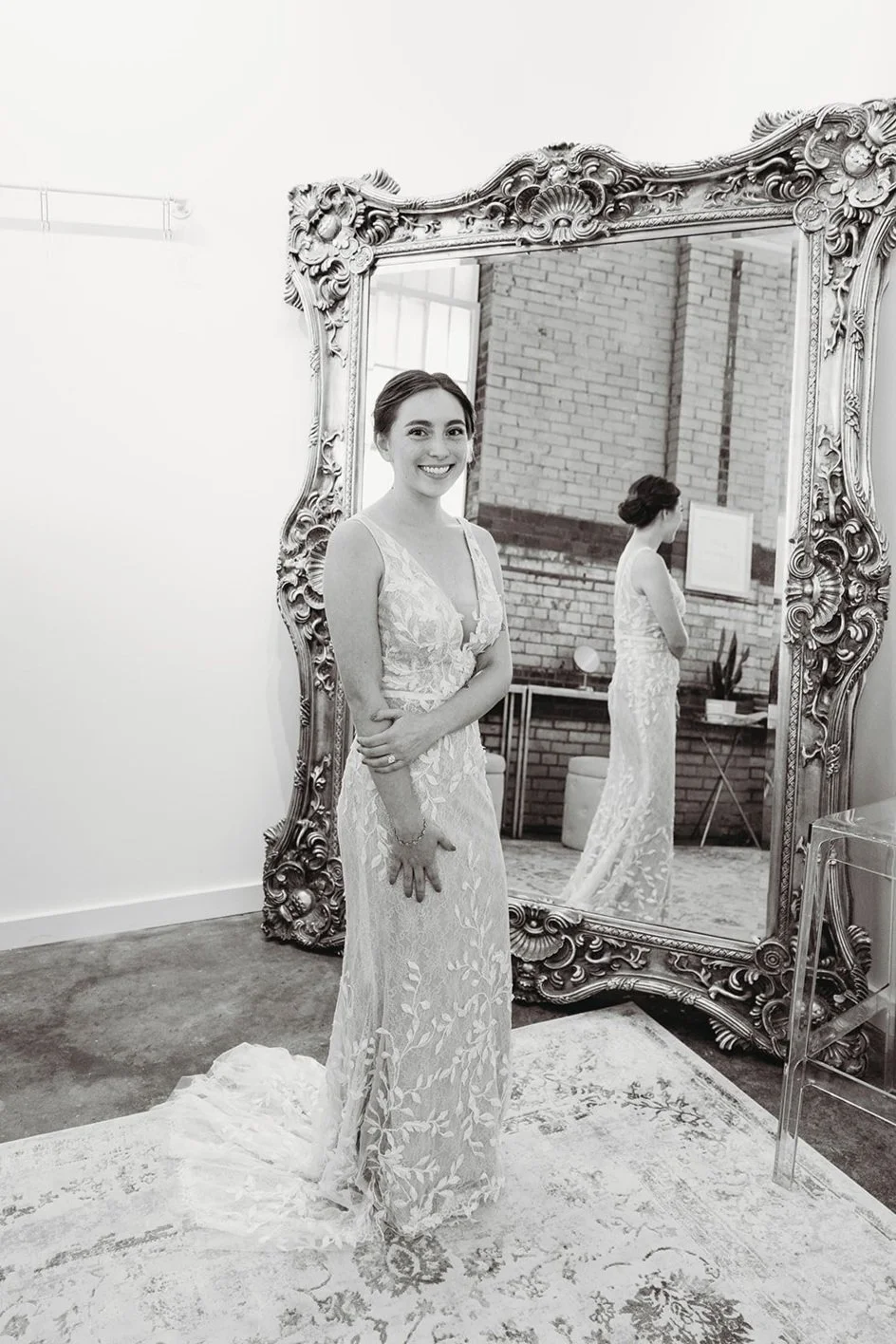 A woman in a wedding dress standing in front of a large ornate mirror, smiling. The mirror reflects her back as she's preparing for her wedding.