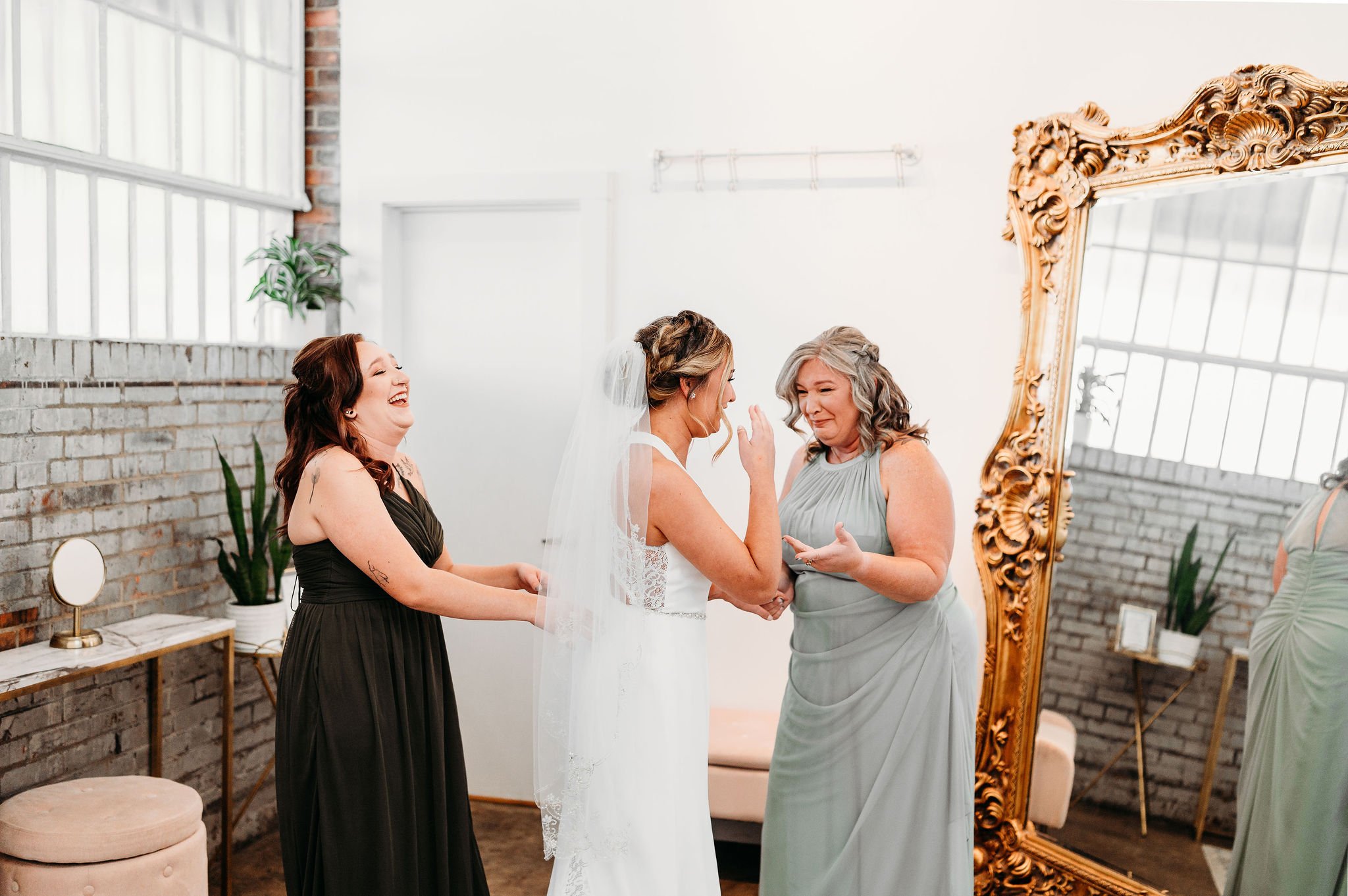 A bride in a white wedding dress and veil is holding hands with an older woman in a light gray dress, while another woman in a black dress laughs nearby. They are in a bright room with a large ornate mirror and a brick wall.