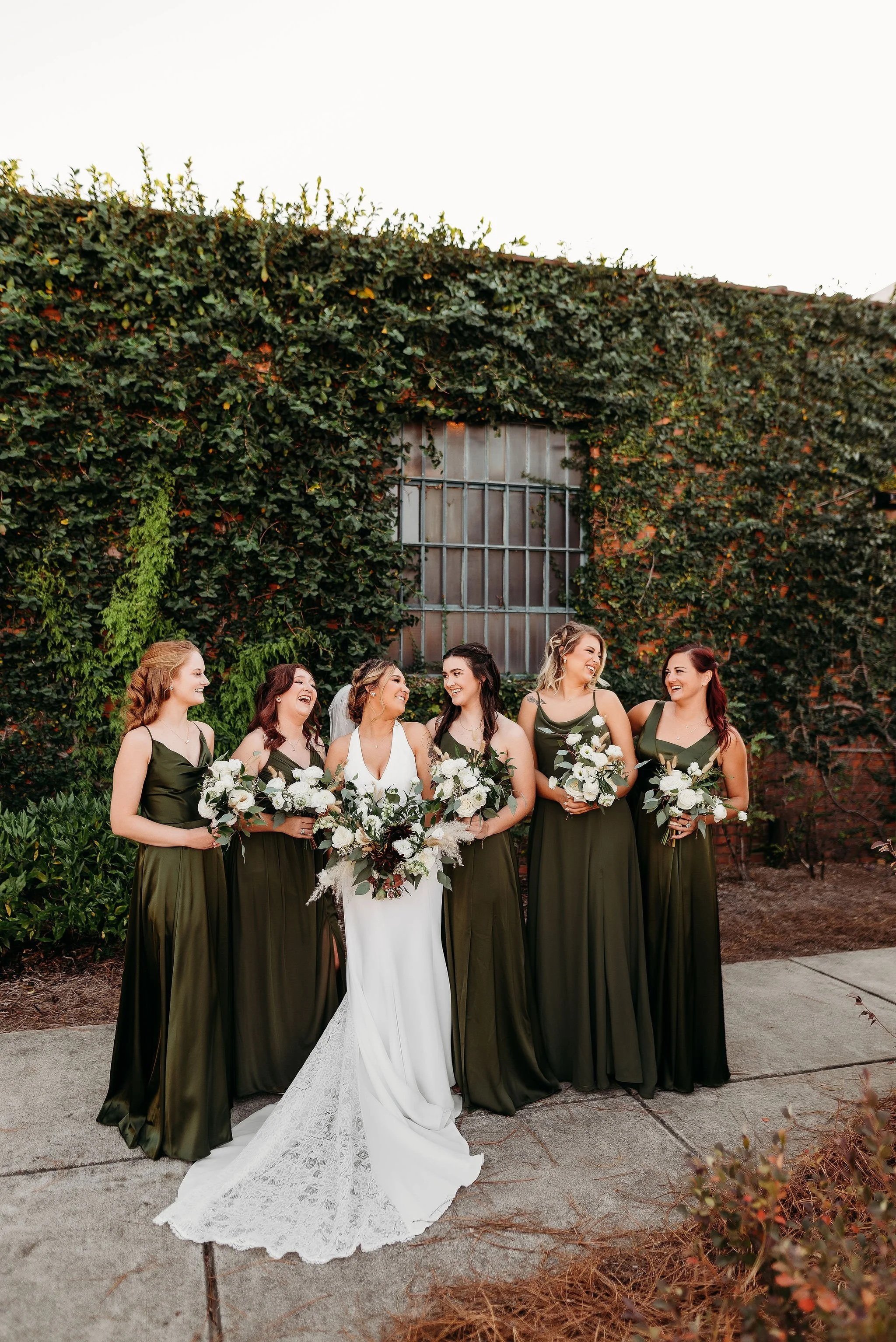 Group of six women, including bride and bridesmaids, standing outdoors in front of a brick wall with green ivy, smiling and holding bouquets, during a wedding celebration.
