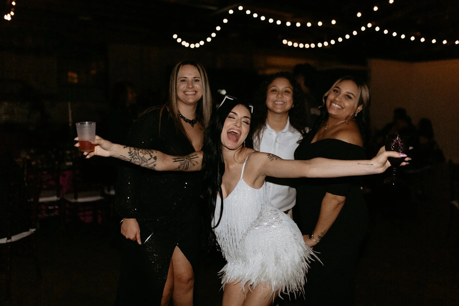 Group of four women celebrating at a party, with string lights overhead, smiling, with one woman in a white dress extending her arms, holding a drink in each hand.