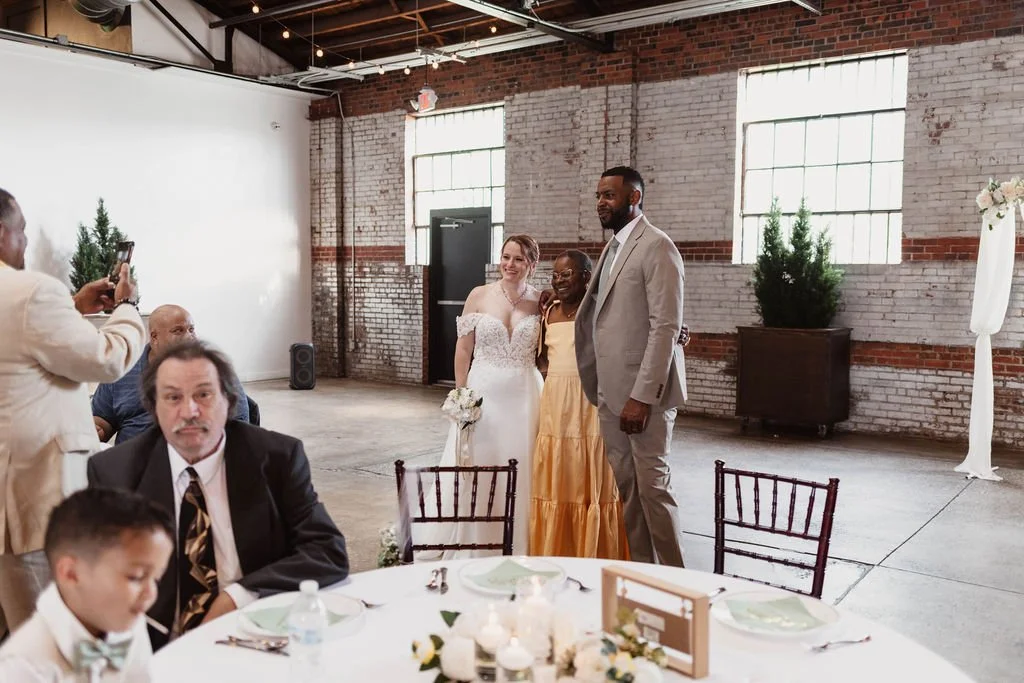 A group of people at a wedding reception, with three individuals standing posing for a photo, including a bride in a white gown and two guests dressed in formal attire, inside a rustic venue with exposed brick walls and large windows.