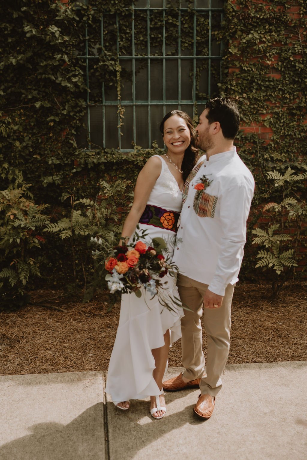 A bride and groom standing outdoors with the bride smiling and holding a bouquet of flowers. The bride is wearing a white dress with floral embroidery and open-toed heels. The groom is dressed in a white shirt, khaki pants, and brown shoes, giving the groom a kiss on the cheek. Behind them is a brick wall covered in green and brown vines, and a window with metal bars.