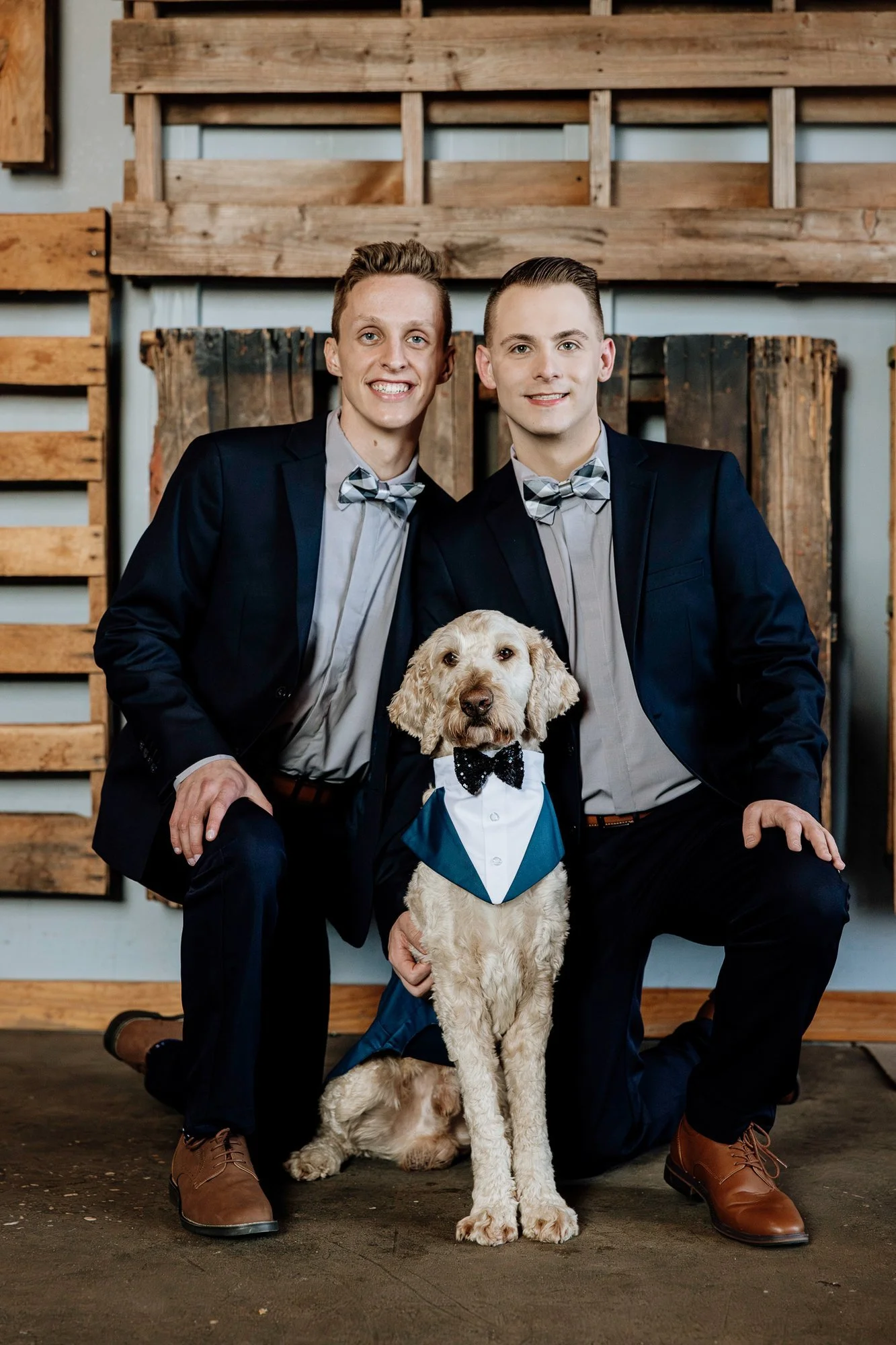 Two men in suits with bow ties and a dog dressed in a tuxedo. They are posing indoors with wooden decor in the background.