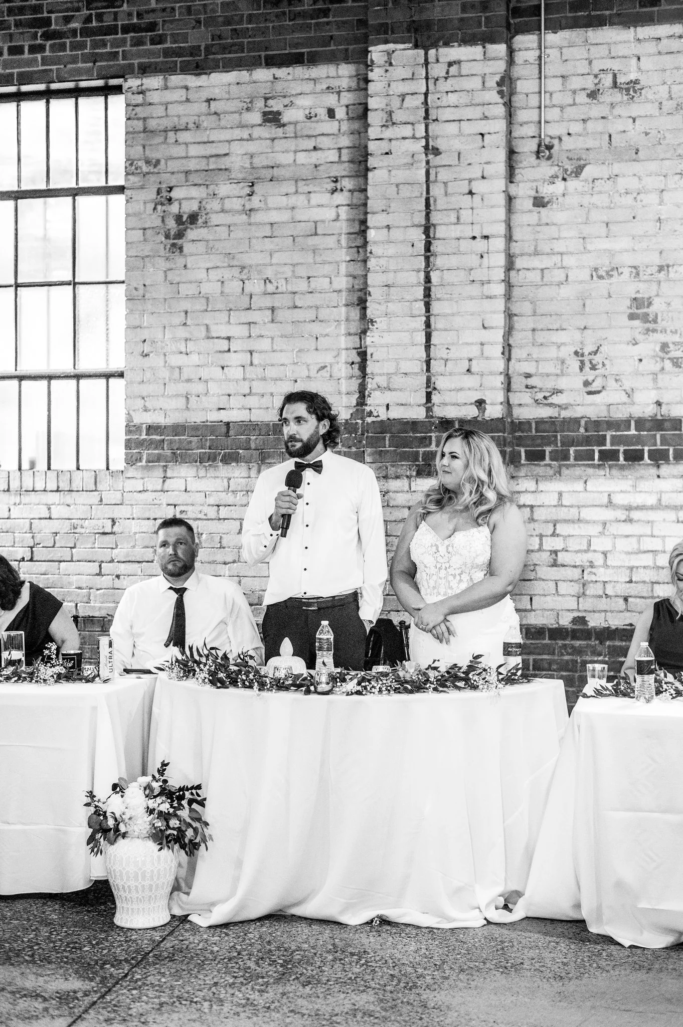 A man in a tuxedo with a bow tie is speaking into a microphone at a wedding reception. A woman in a white dress stands beside him. They are at a decorated head table with floral arrangements, water bottles, and glasses, against a brick wall backgroun