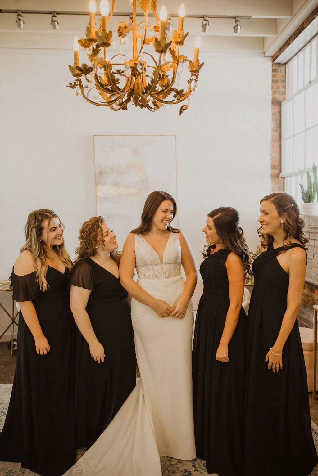 A bride in a white wedding dress smiling and talking with five bridesmaids dressed in black gowns in a decorated indoor setting with a chandelier hanging from the ceiling.