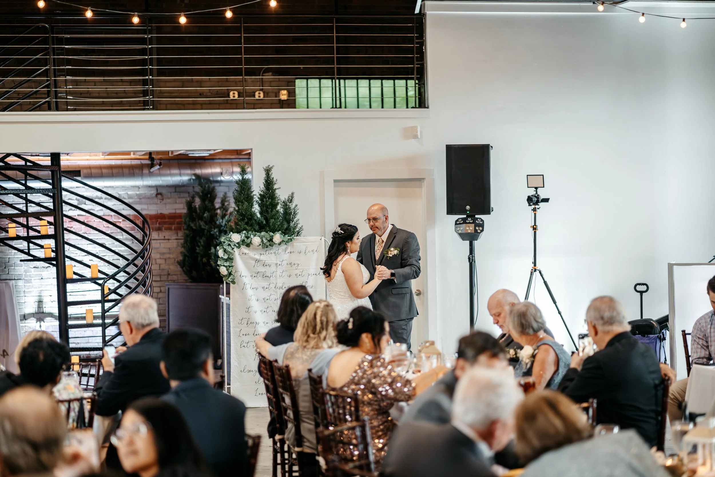 A bride and groom dancing at their wedding reception, surrounded by seated guests, in a modern indoor venue with string lights and greenery decorations.