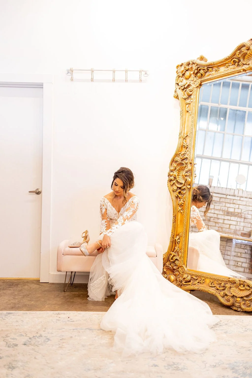 Bride in a white wedding dress sitting on a pink bench, putting on her wedding shoes, in front of a large ornate gold mirror in a bright, minimalist room.