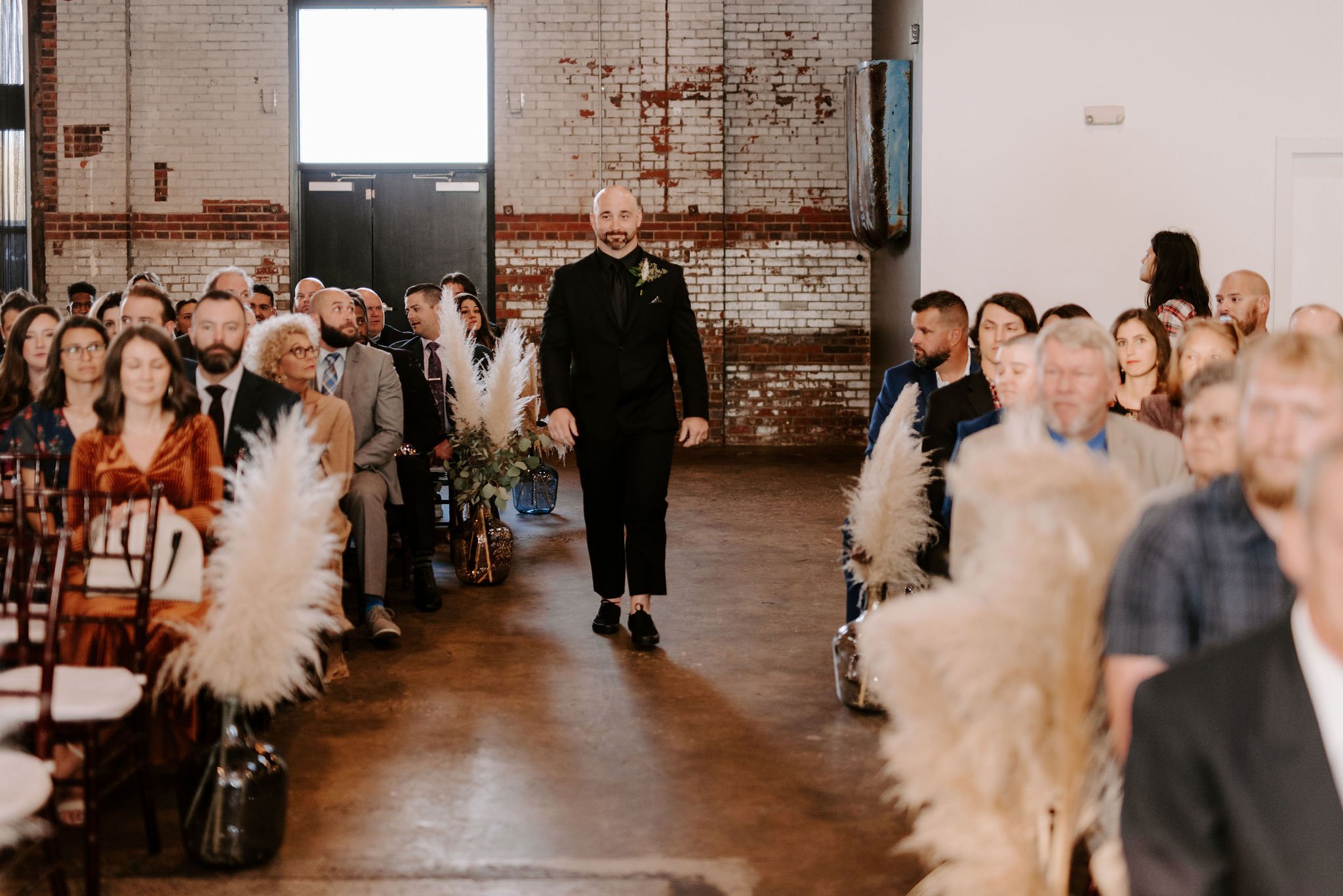 Man in a black suit walking down the aisle at a wedding in an industrial-style venue with exposed brick walls and large windows, guests seated on either side.