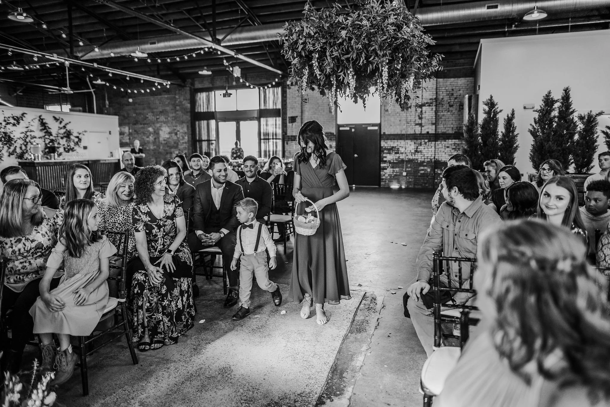 A black-and-white photo of a woman holding a basket, walking down the aisle in a venue with seated guests on either side, likely at a wedding or special event.