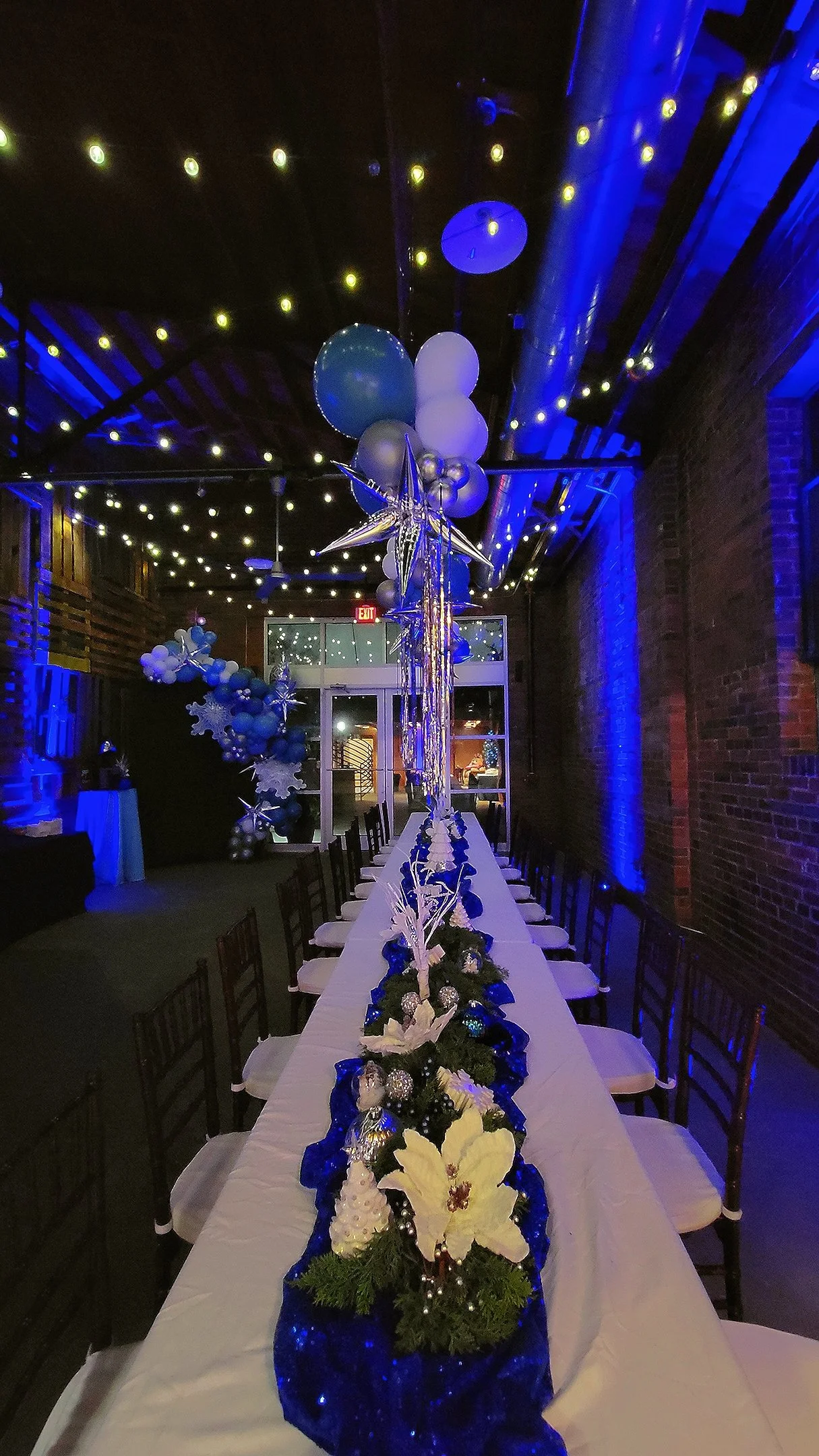 Long banquet table decorated with Christmas flowers, ornaments, and blue fabric runner, with balloon and star-shaped decorations hanging from the ceiling at a holiday party venue with blue and warm lighting.