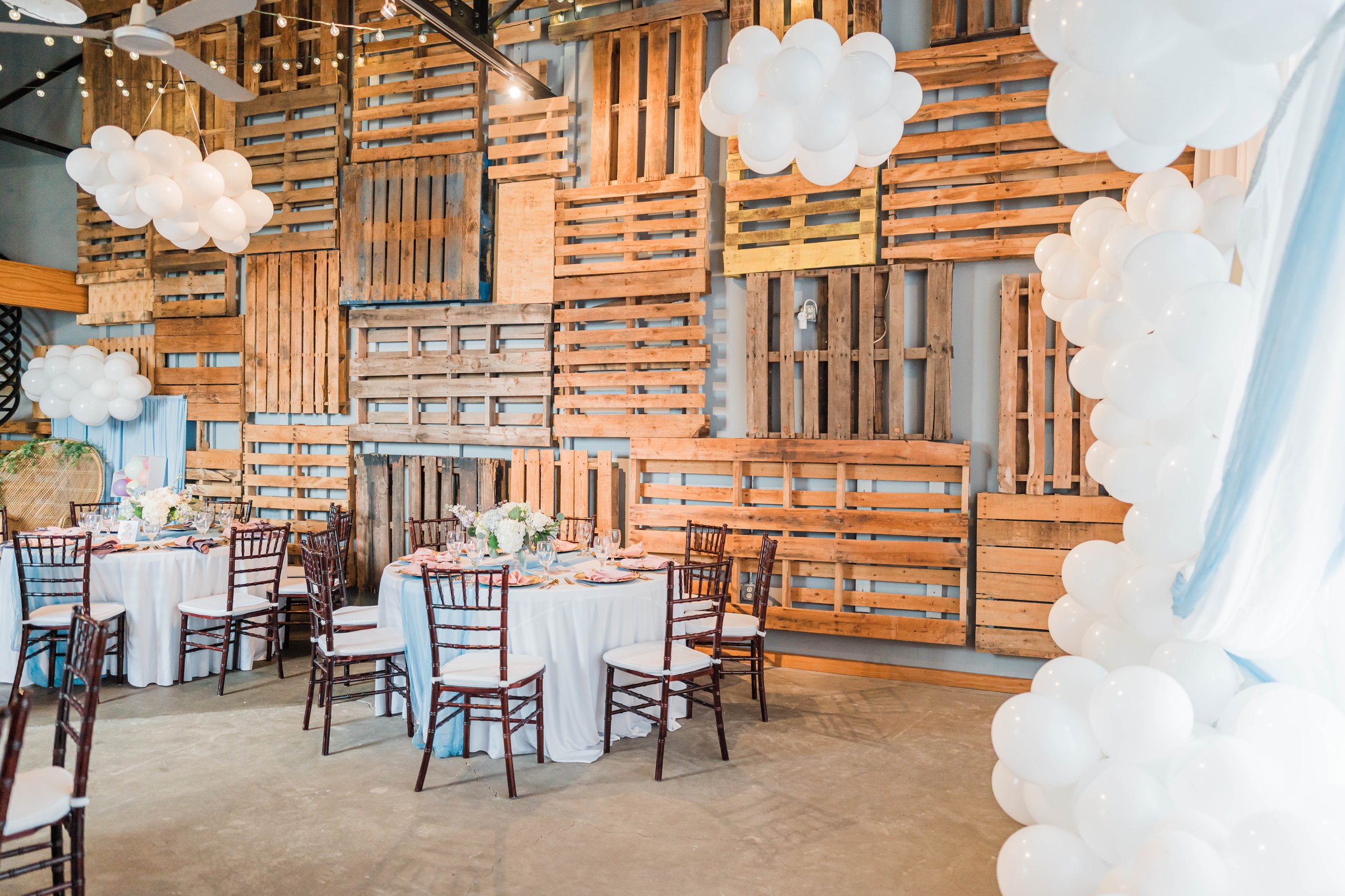 Indoor event space decorated with white balloons and reclaimed wood pallet wall, with round tables draped in white tablecloths, floral centerpieces, and wooden chairs.