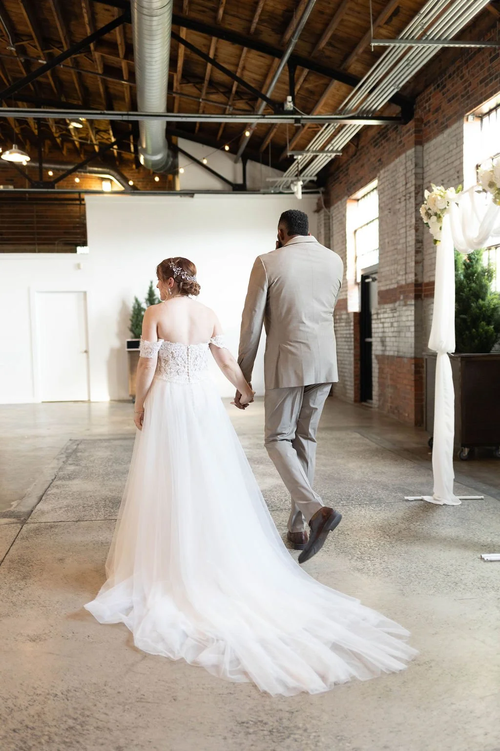 A bride and groom holding hands walking away in a rustic venue with exposed brick walls and high wooden ceiling.