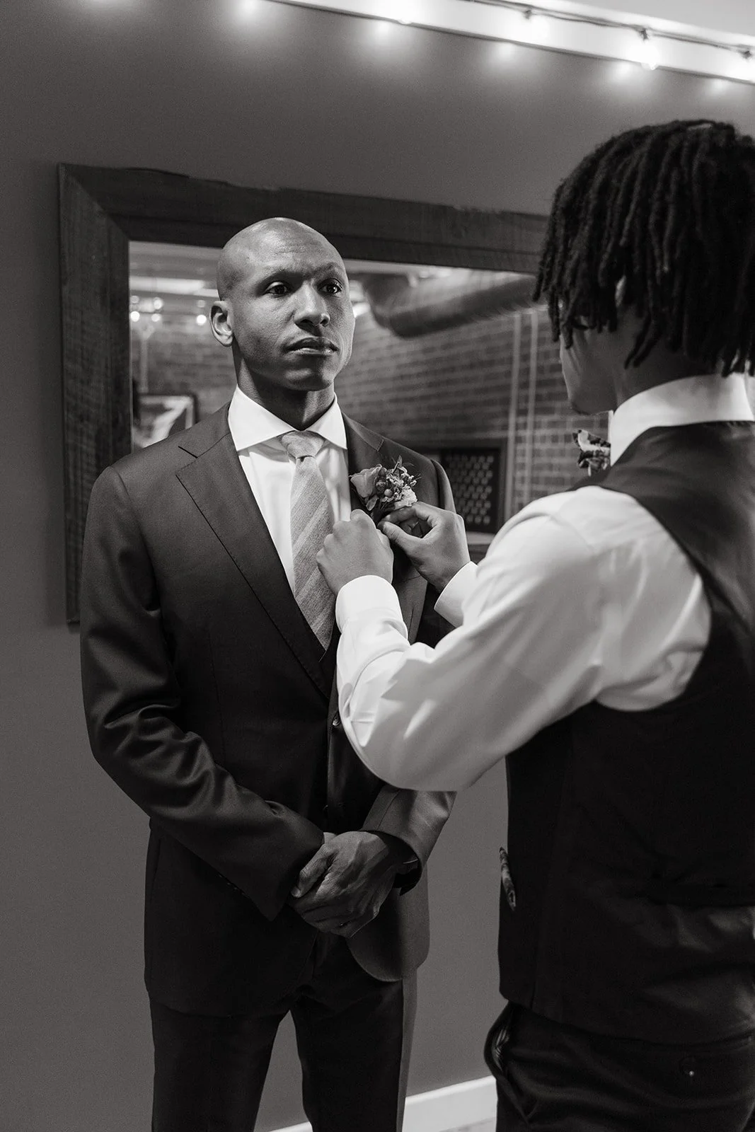 A man in a suit standing in front of a mirror while another man helps him adjust a boutonniere on his lapel.