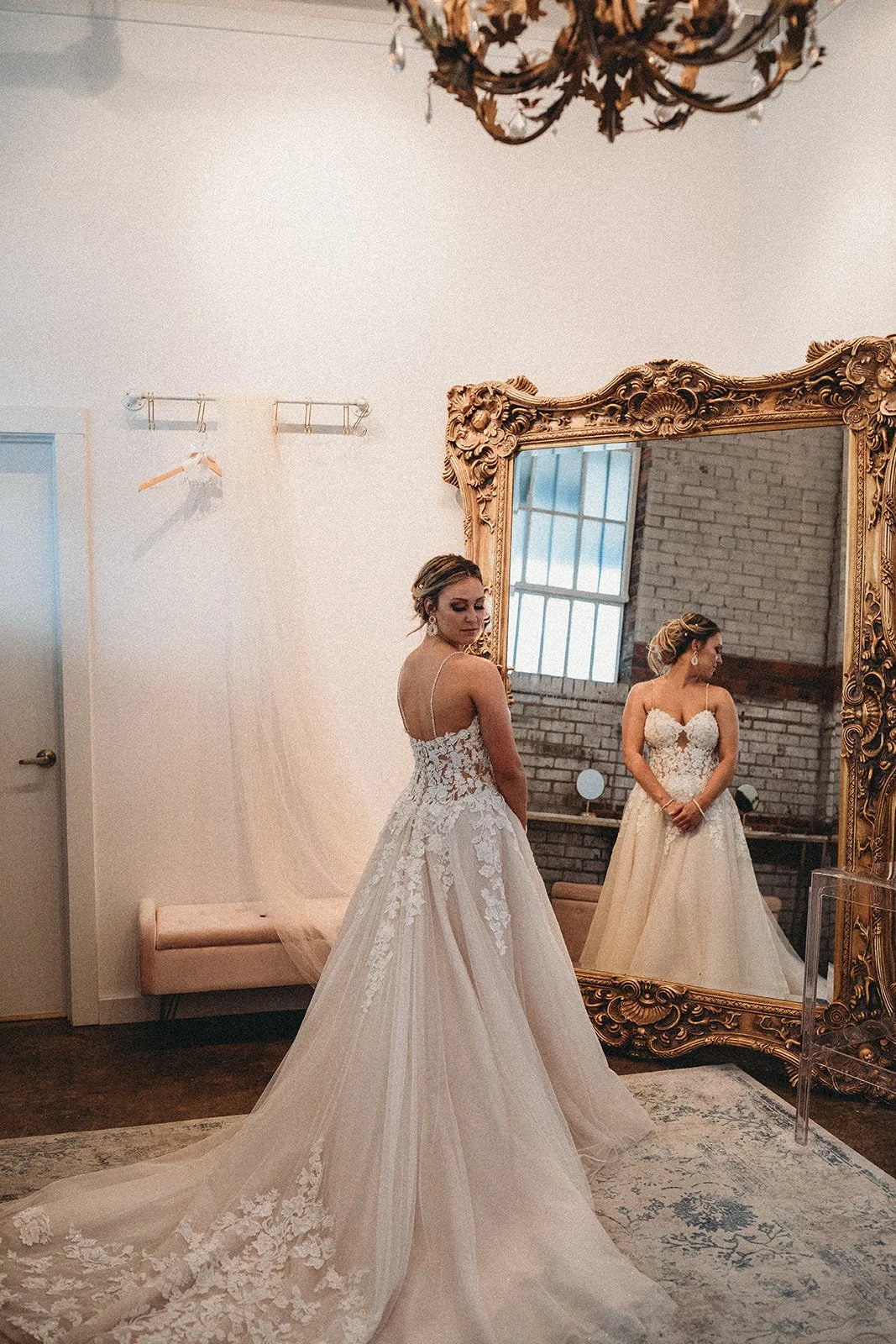 A bride trying on a wedding dress in a fitting room, looking at her reflection in an ornate gold-framed mirror.