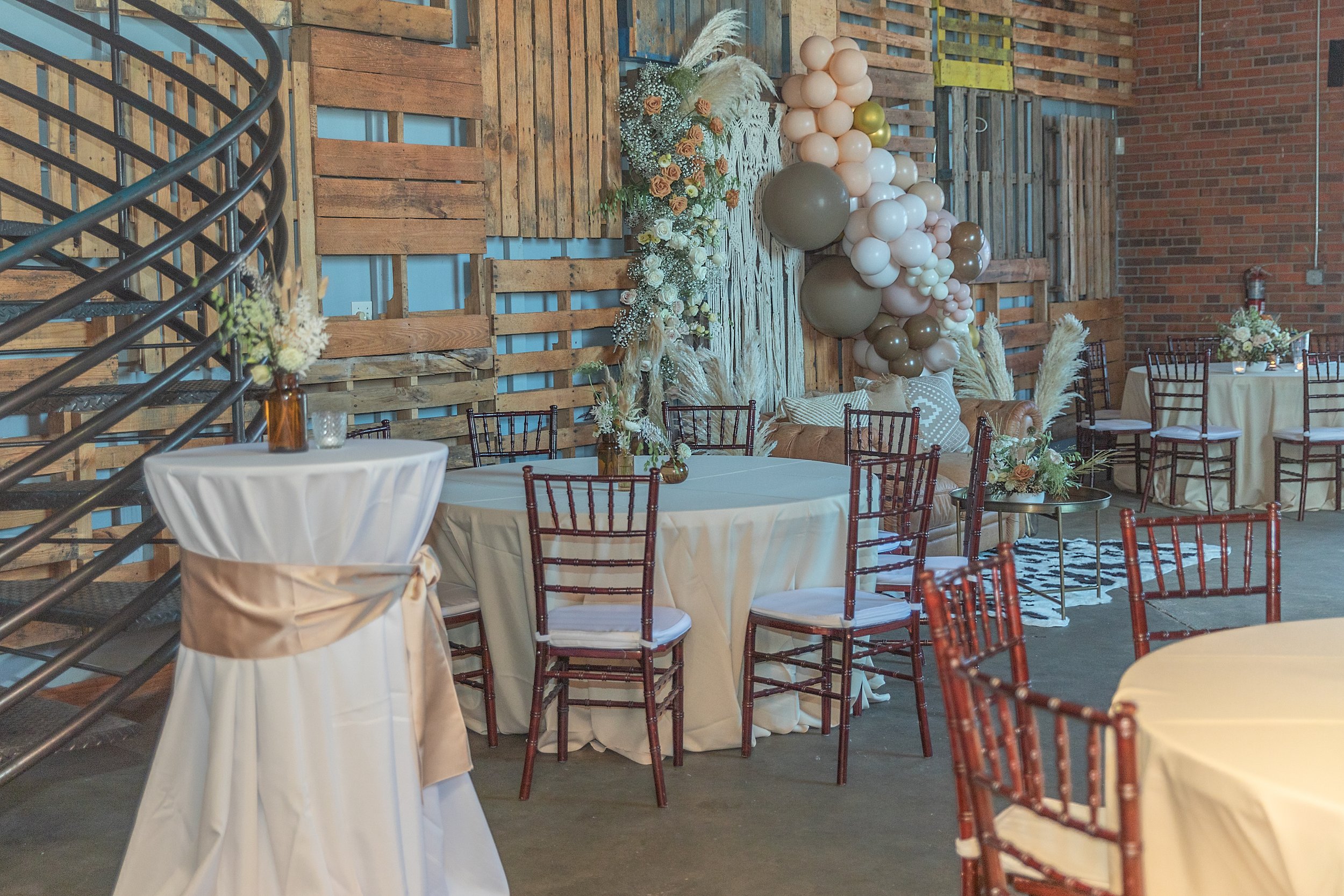 Event space decorated with balloon arrangements, flowers, and draped fabric for a celebration or wedding reception, featuring round tables with white tablecloths and floral centerpieces, rustic wooden wall backdrop, and multiple chairs.