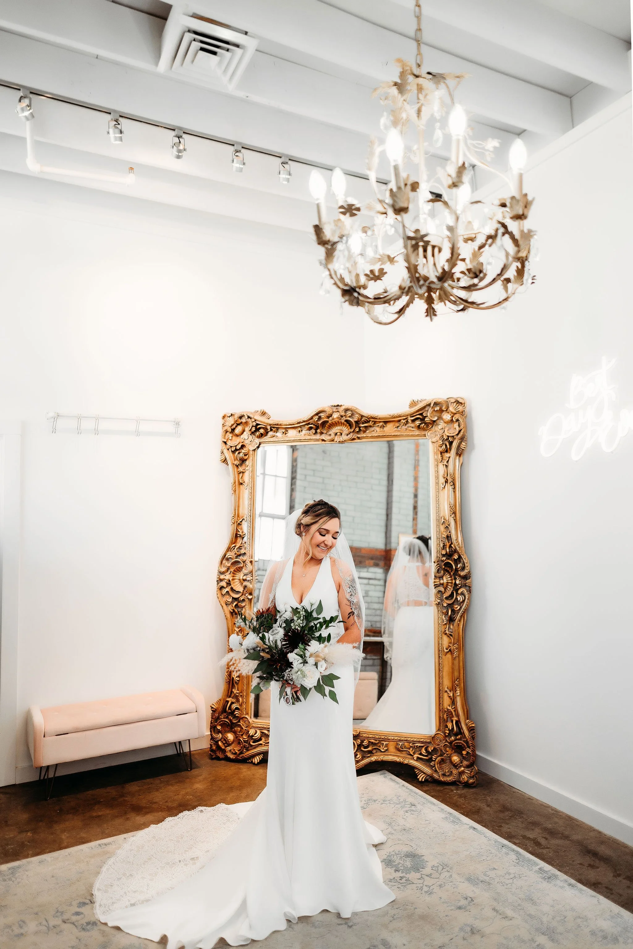 A bride in a white wedding dress holding a bouquet of flowers, standing in front of a large ornate mirror with a gold frame, in a room with white walls and a chandelier hanging from the ceiling.