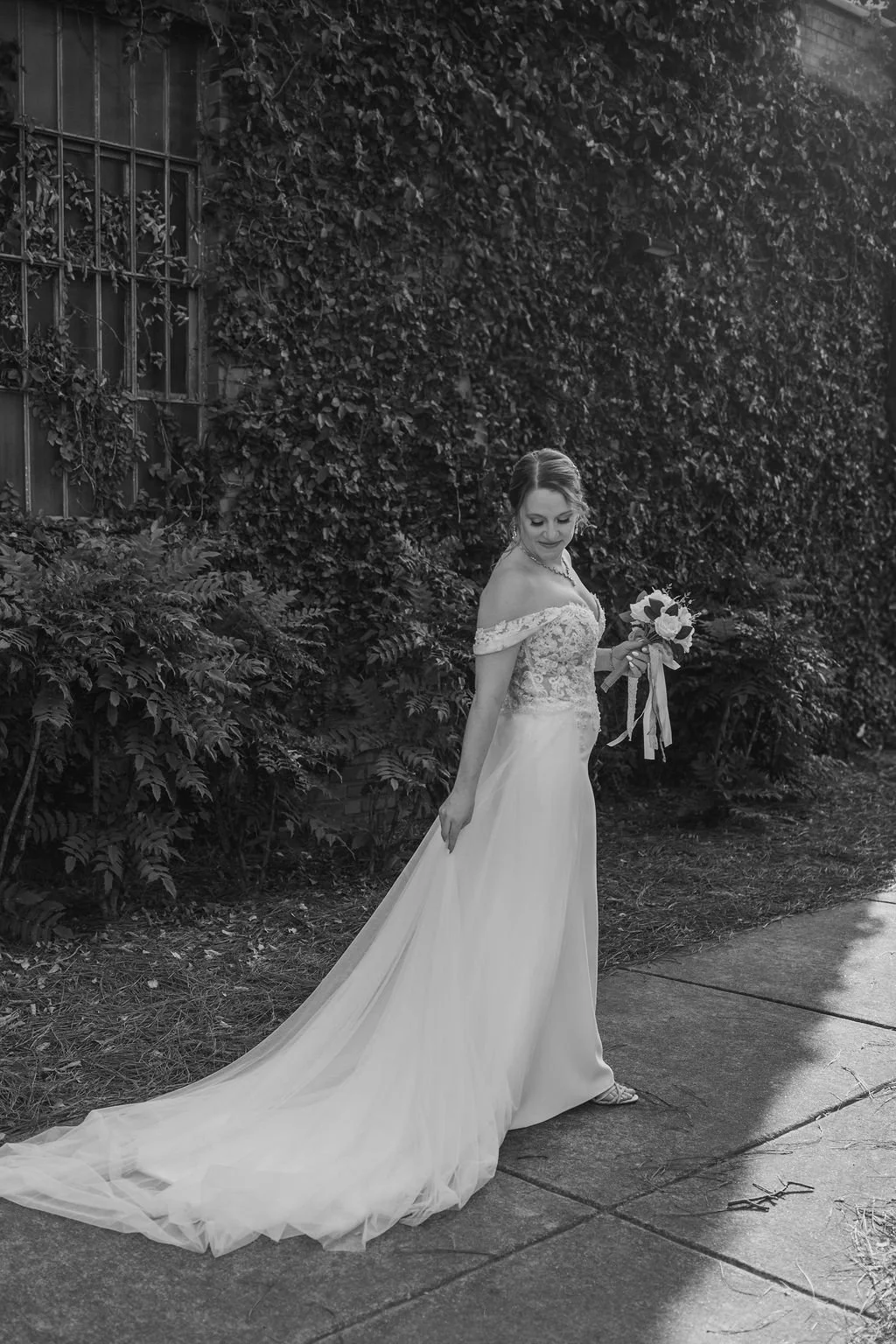 A bride in an off-shoulder wedding dress holding a bouquet of flowers, standing on a sidewalk next to a wall covered with ivy, looking down and smiling.