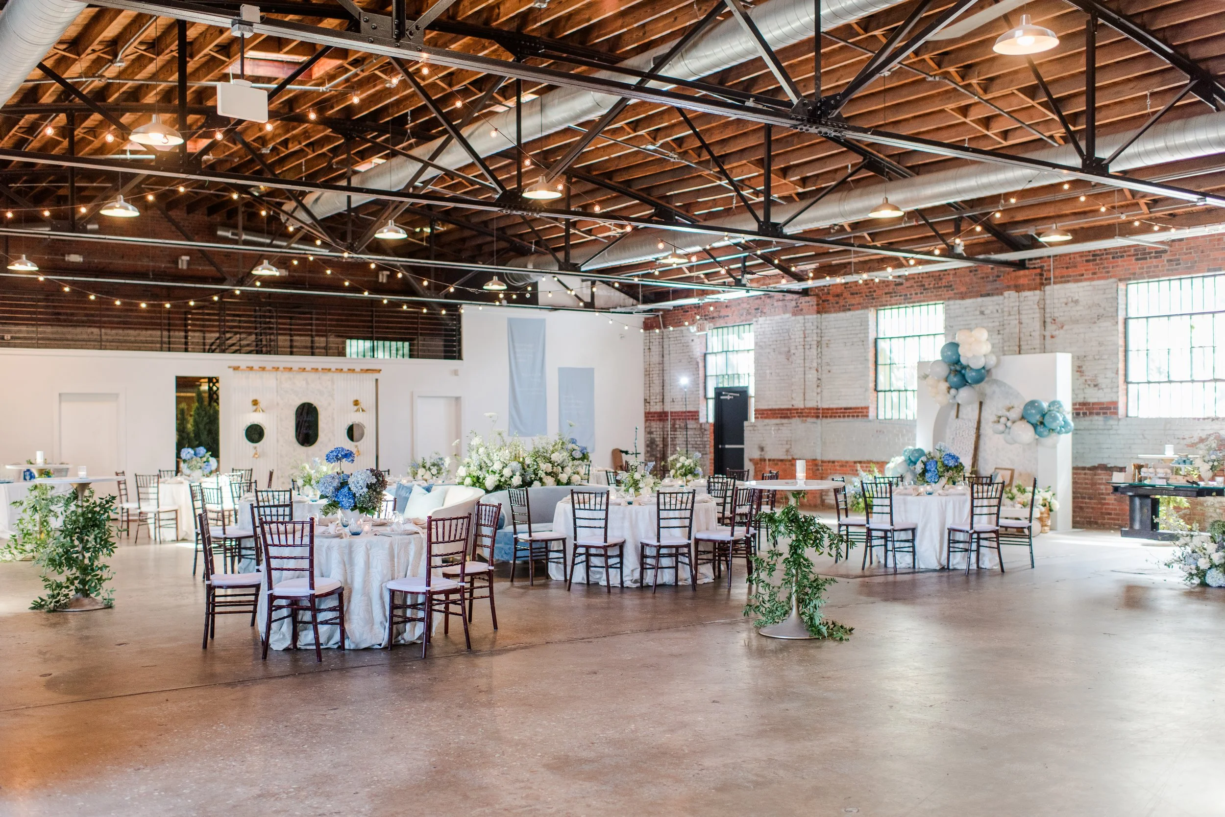 Interior of a decorated event hall with round tables covered with white tablecloths, floral centerpieces, wooden chairs, balloon arrangements, and string lights hanging from a wooden ceiling with exposed beams.