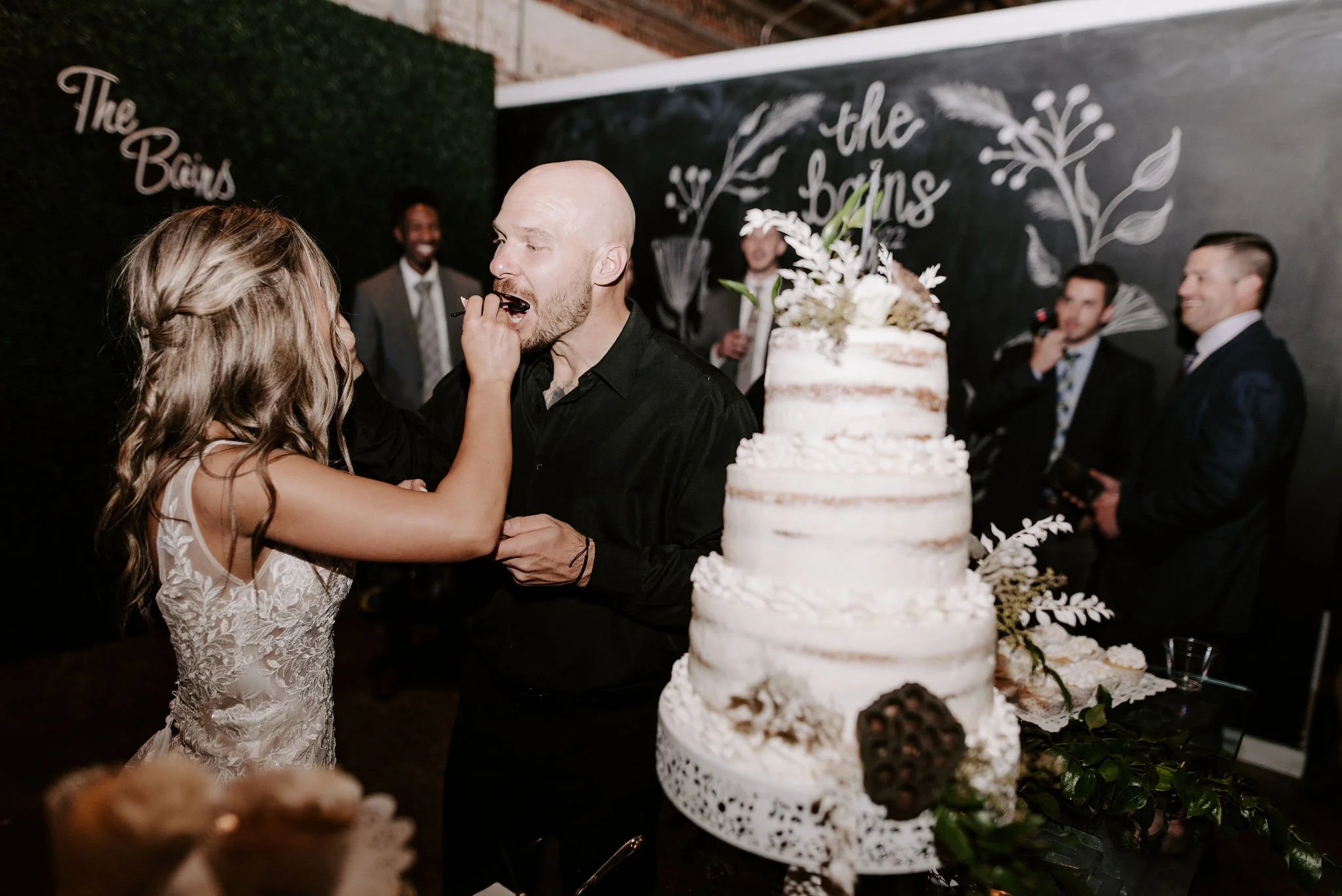 A bride in a lace wedding dress feeding cake to a groom in a black shirt during a wedding reception, with a tall layered wedding cake decorated with flowers and leaves in the foreground, and a dark wall with decorative writing and guests in tuxedos i