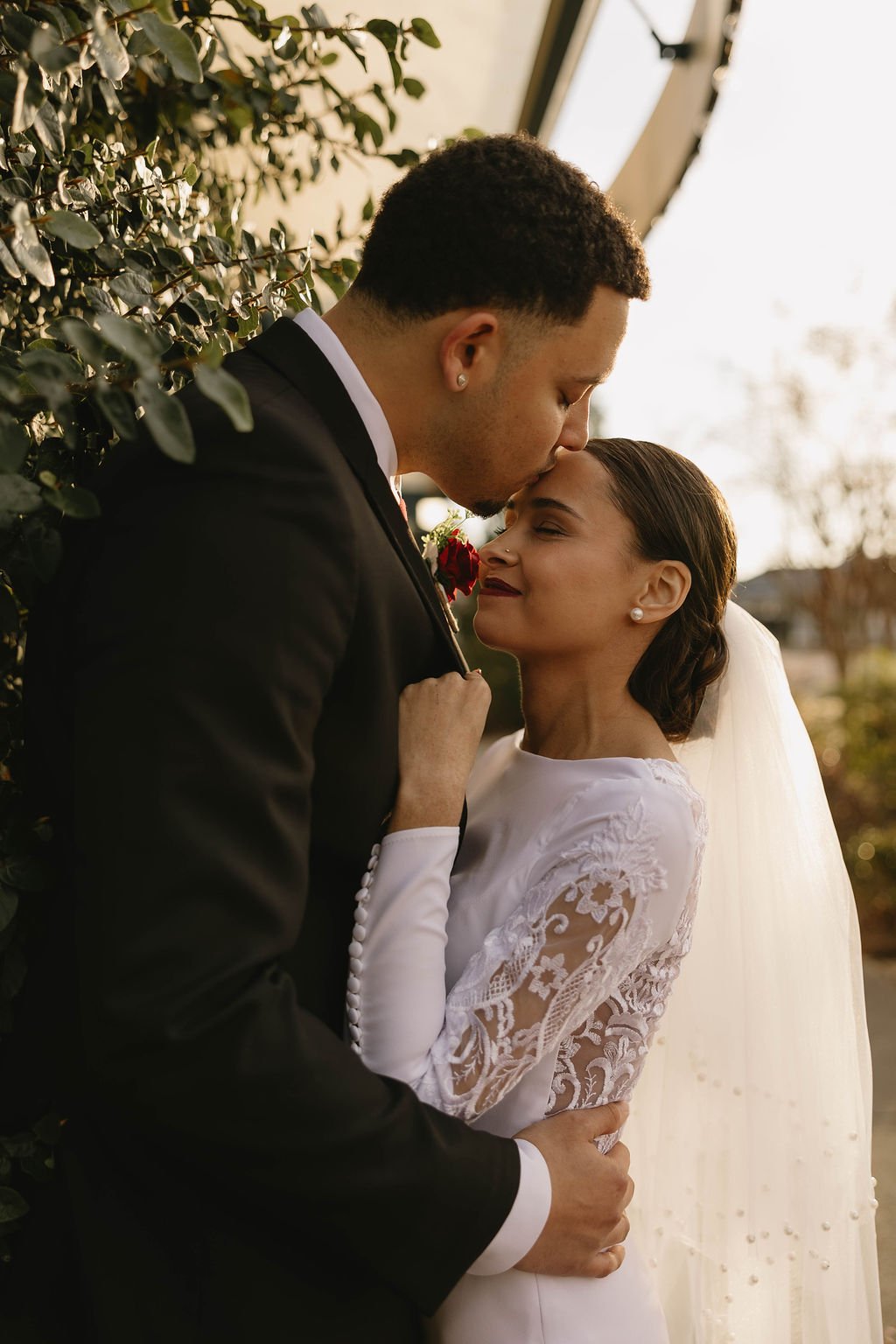 A bride and groom sharing a tender kiss outdoors, with the groom pressing his forehead to the bride's, both eyes closed and smiling gently, during sunset.