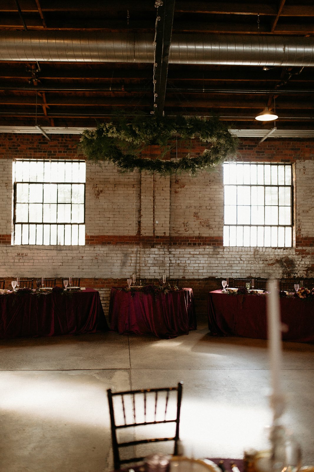 An industrial-style event space with brick walls, large windows, a wooden ceiling with exposed ductwork, and a green wreath hanging from the ceiling. There are tables with burgundy tablecloths set with glasses and plates along the wall.