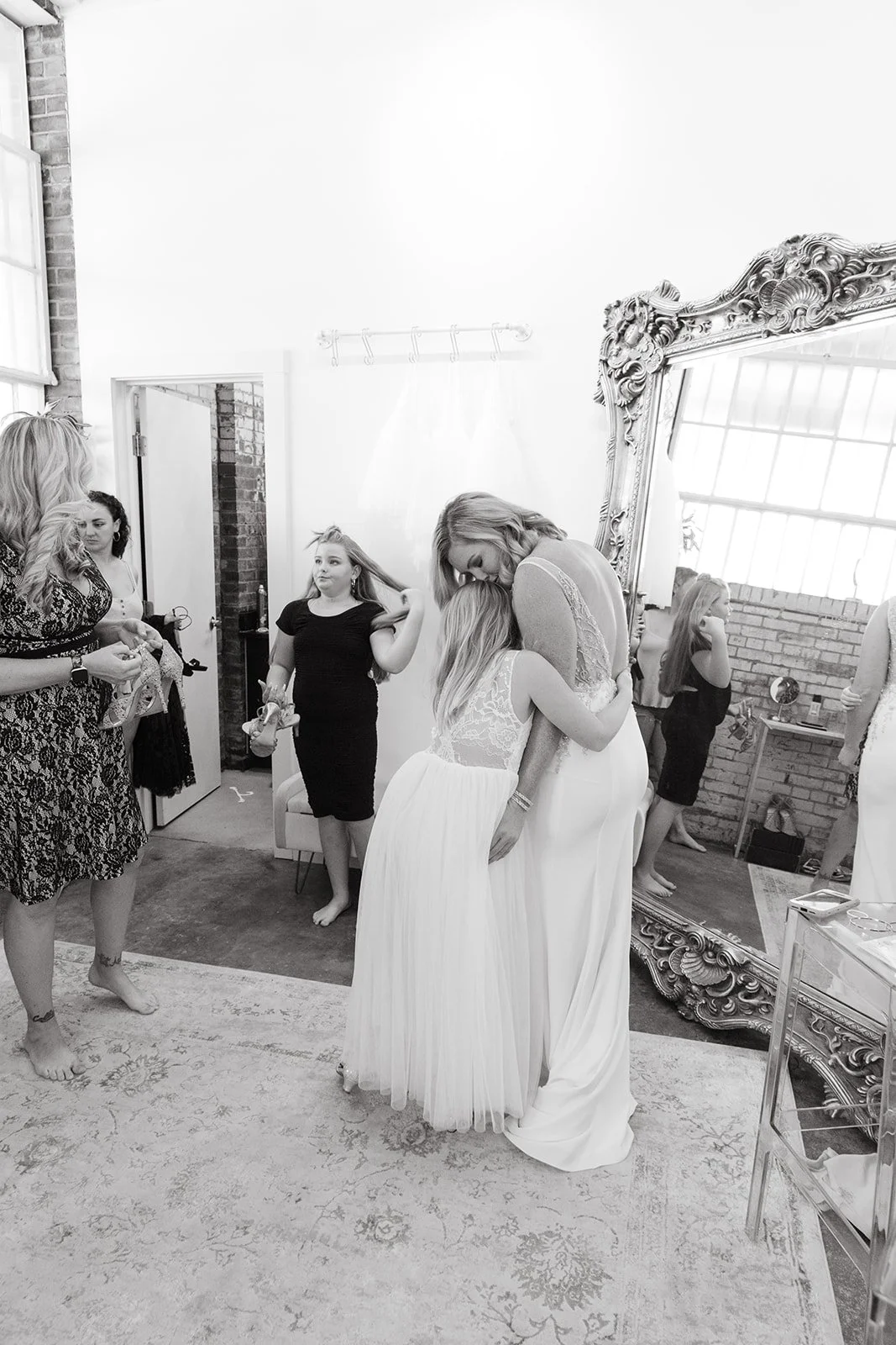 A bride in a white wedding gown hugging a young girl in a similar dress inside a bridal studio, with other women in the background, some holding gifts or accessories.