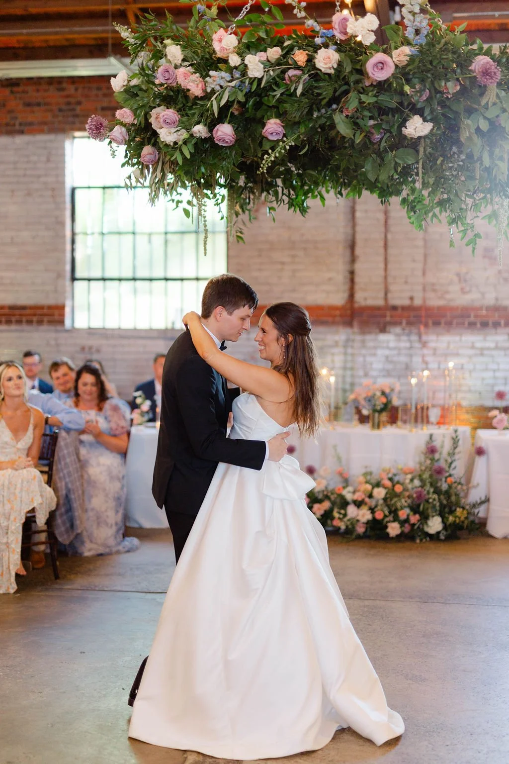 A bride and groom dance during their wedding reception, with guests seated at tables decorated with flowers in the background and a large floral arrangement hanging above them.