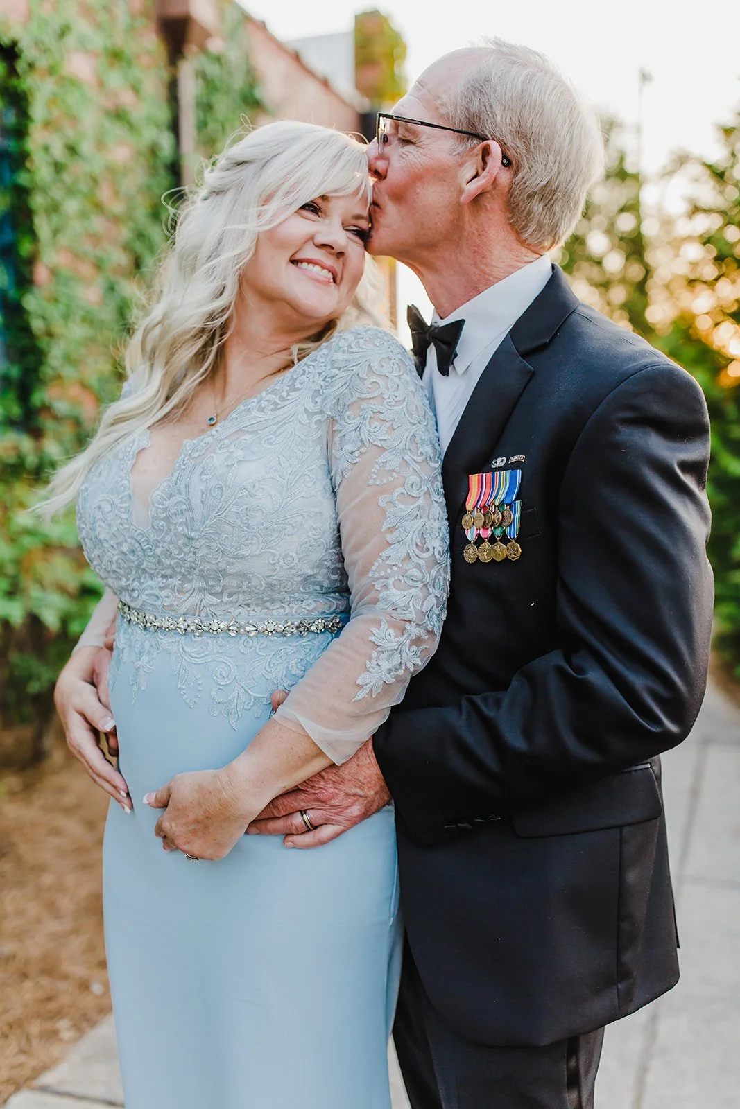 An elderly man in a military uniform with medals kisses a smiling woman in a light blue lace dress. They are outdoors with greenery and a brick wall, during sunset.