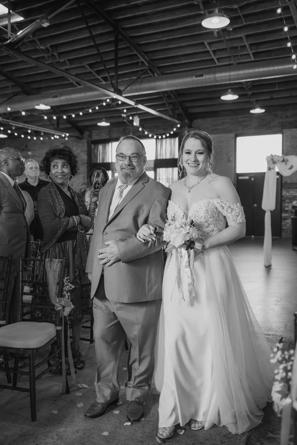 A bride walking down the aisle with her father at her wedding ceremony in an industrial-style venue with string lights.
