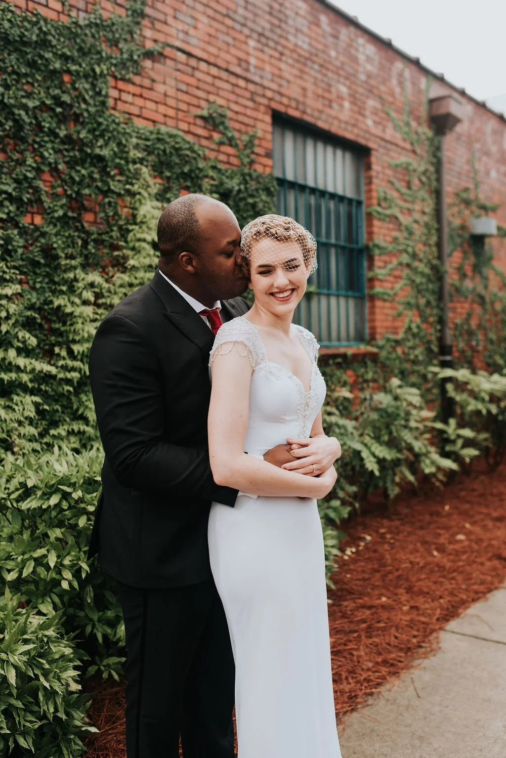 Bride and groom posing in front of the signature green ivy wall at Studio 215 in downtown Fayetteville, NC; a lush urban garden photography backdrop at our industrial warehouse venue near Fort Bragg and Raleigh.