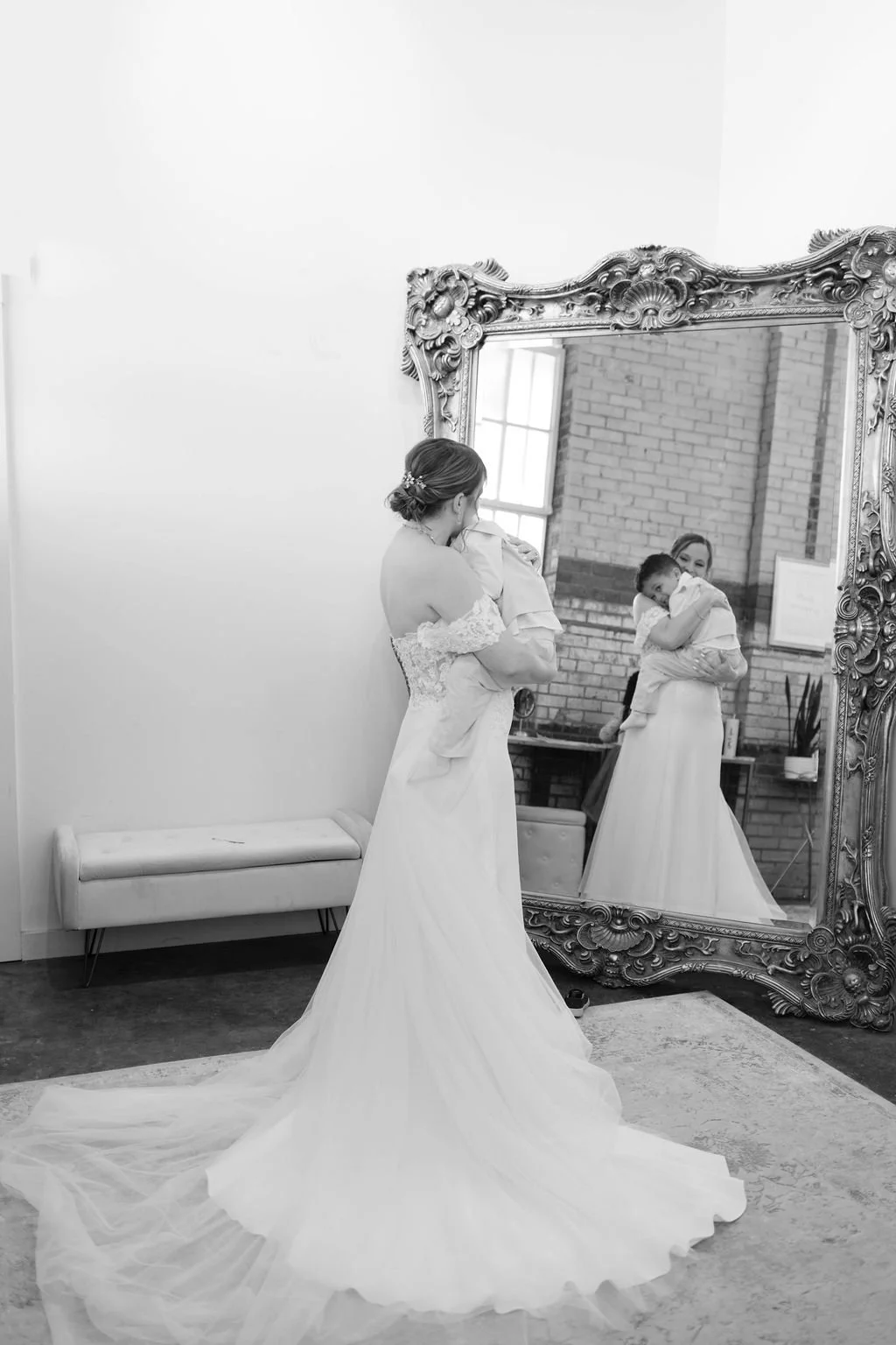 A bride in a wedding dress is admiring herself in a large ornate mirror, holding a child on her shoulder, in a room with brick walls and a window.
