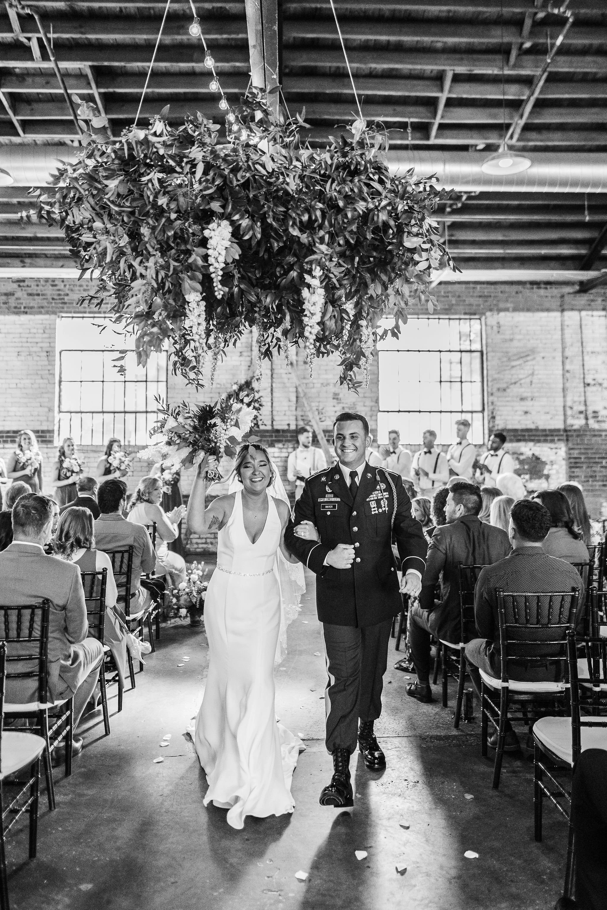 A bride and a groom, a military soldier, walking down the aisle after their wedding ceremony in an industrial-style venue with exposed brick walls and large windows, surrounded by seated guests and wedding party in the background.