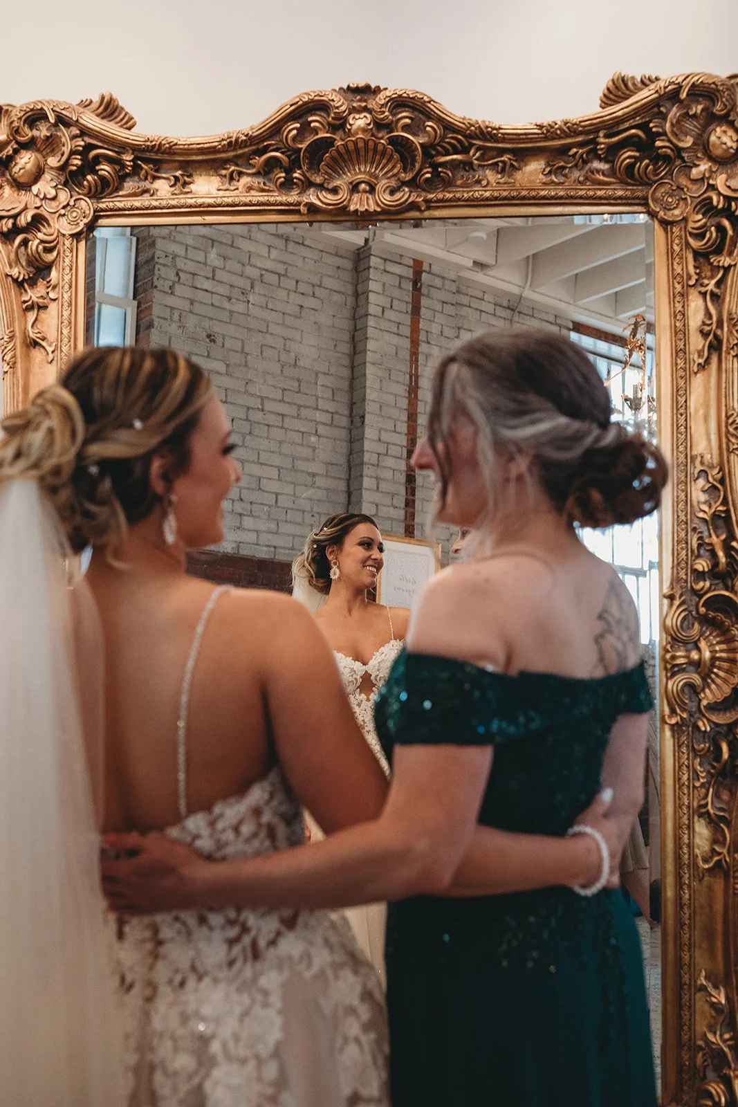 A bride and another woman are smiling and holding each other while looking into a large ornate gold mirror in a bridal boutique. The bride is wearing a lace wedding gown, and the woman is dressed in a dark green off-the-shoulder dress with sequins.