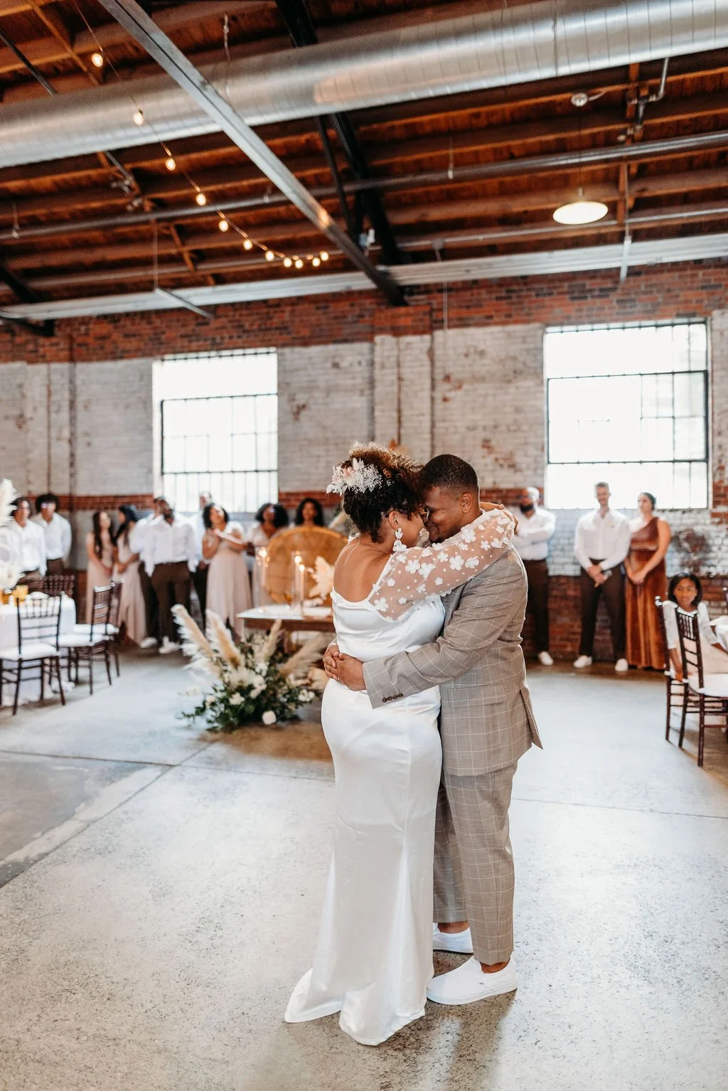 A bride and groom dancing closely at their wedding reception, surrounded by guests in a rustic industrial-style venue with brick walls and large windows.