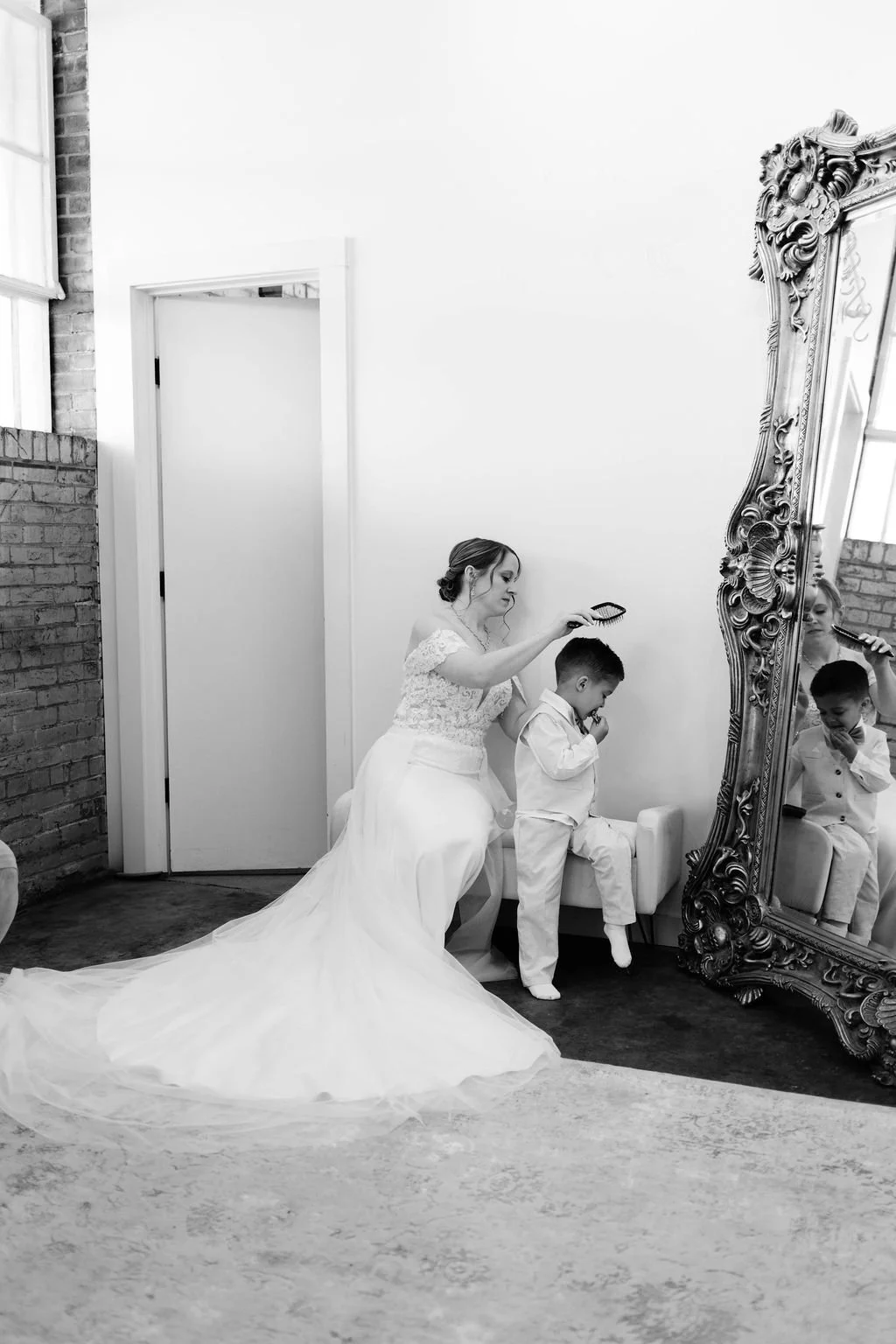 A bride in a wedding dress combing a young boy's hair in front of a large ornate mirror, with both looking at their reflection.