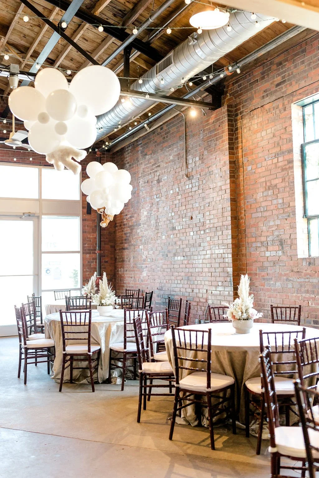 ndustrial chic event space at Studio 215 in Fayetteville, NC, featuring exposed brick walls, hanging cloud balloon installations with teddy bears, and elegant table settings with pampas grass centerpieces for a baby shower or wedding reception