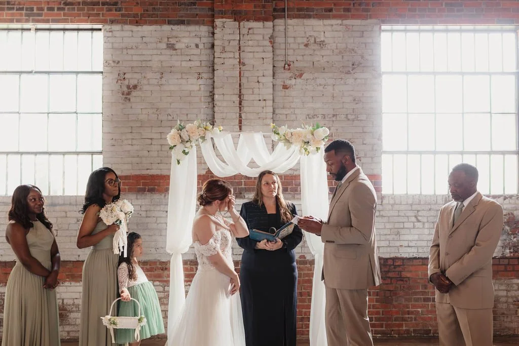 A wedding ceremony with a bride, groom, and officiant in front of a decorated arch, with bridesmaids and a flower girl standing nearby, inside a brick-walled venue.