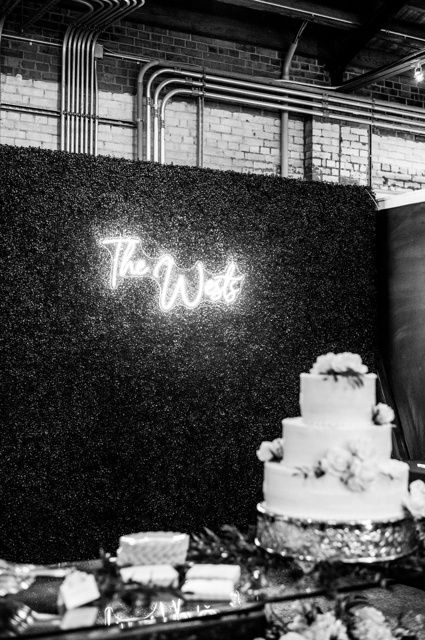 A three-tier wedding cake adorned with flowers on a reflective silver cake stand, set in front of a black textured wall with a neon sign reading 'The Wets' in cursive, and exposed metal pipes and brick wall in the background.