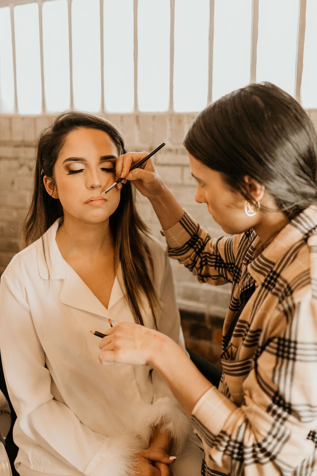 Makeup artist applying eyeshadow to a woman with closed eyes in a well-lit room.
