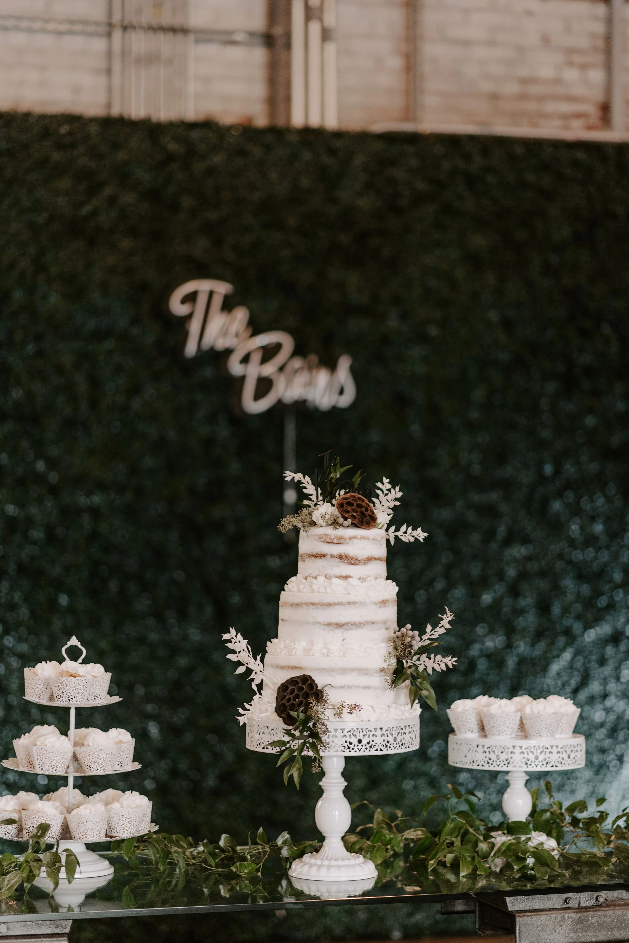 A three-tier naked wedding cake decorated with sprigs of greenery and brown flowers, placed on a white cake stand. Cupcakes with white frosting are arranged on white stands on either side. A green leafy garland decorates the table. In the background,