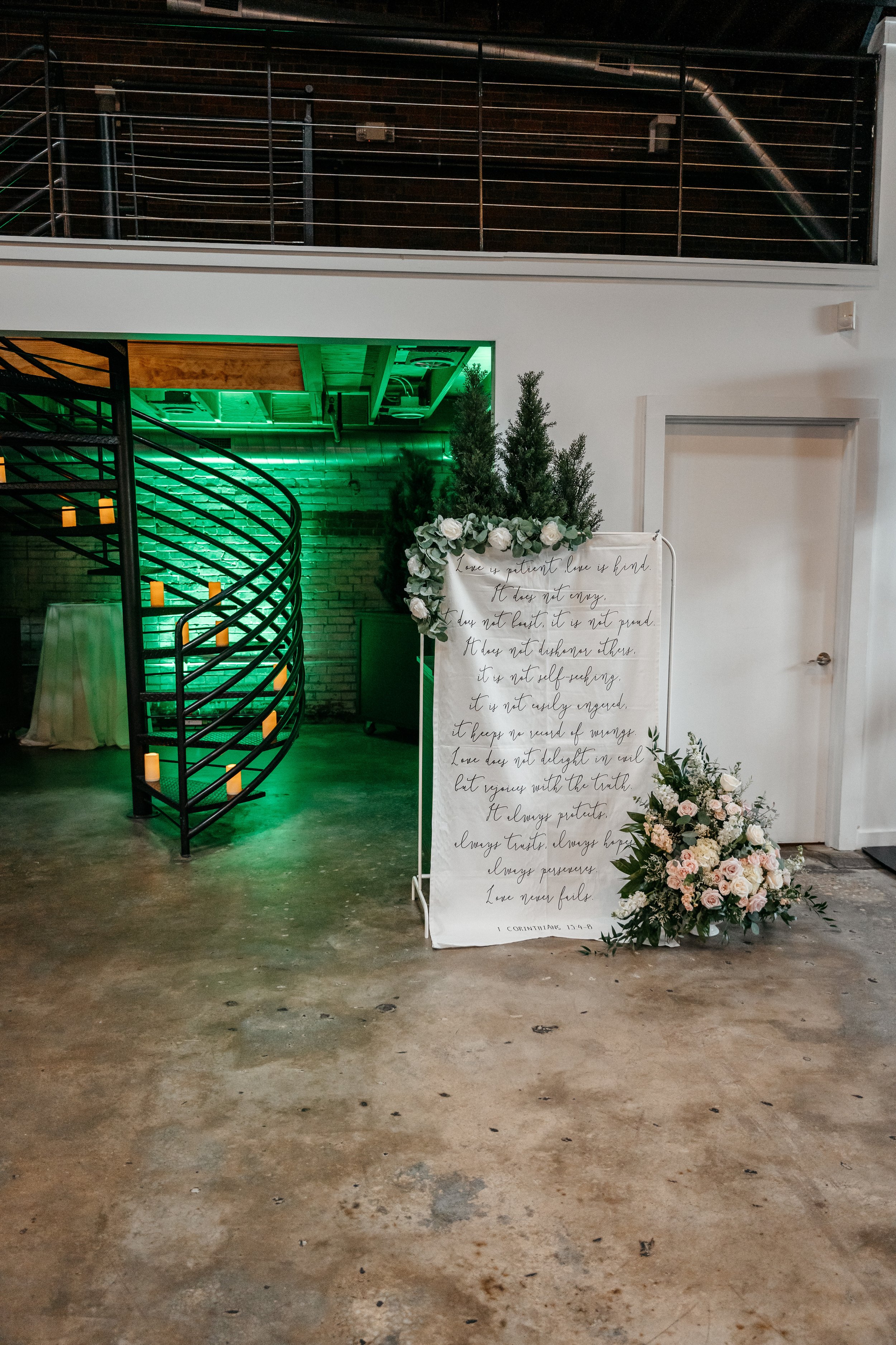 Wedding or event sign with a floral arrangement and greenery, featuring a handwritten quote from 1 Corinthians 13:4-8, in an indoor venue with a black spiral staircase and green accent lighting.