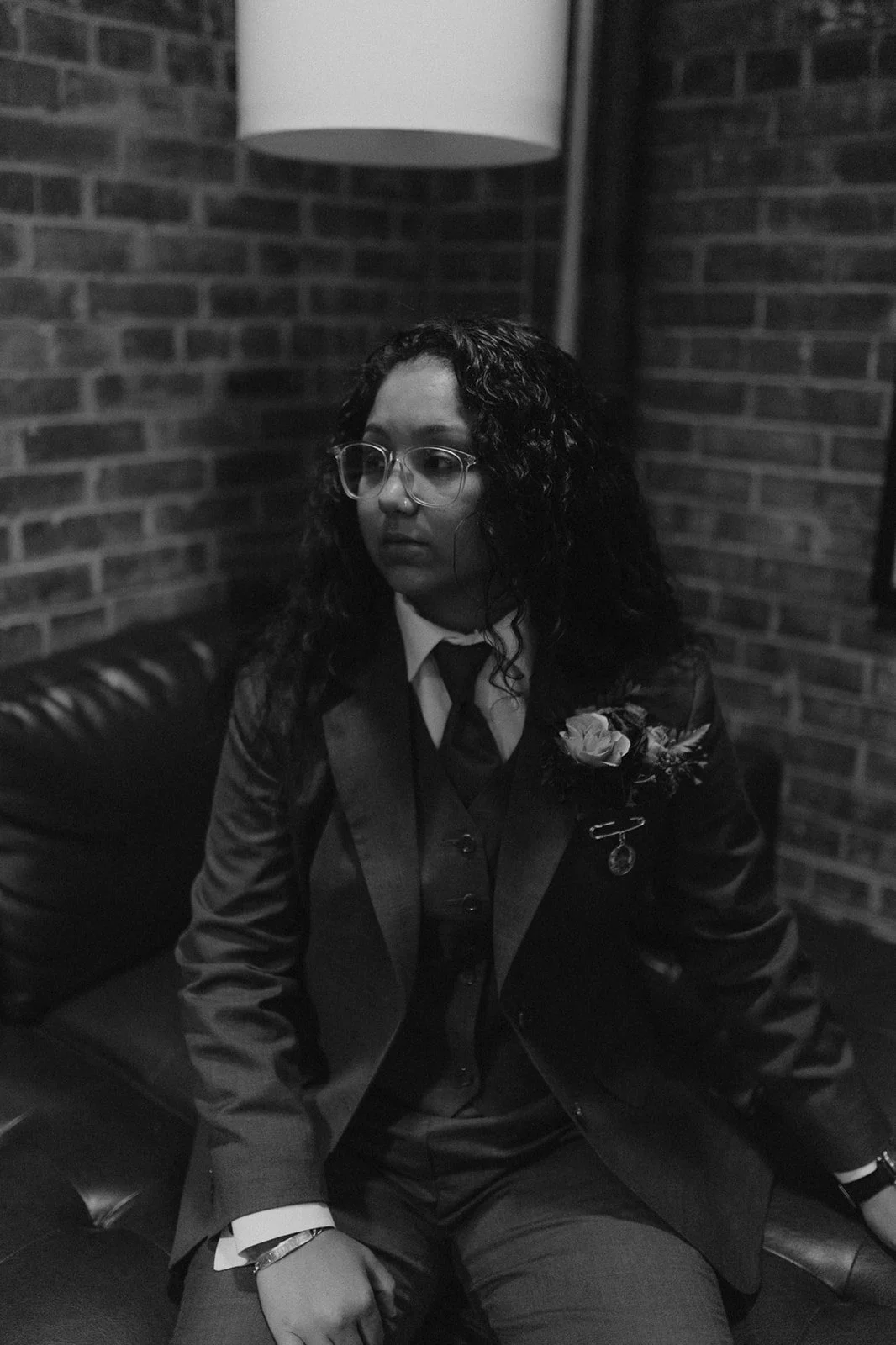 A woman with curly dark hair, glasses, and a serious expression, dressed in a formal suit with a boutonnière, sitting on a leather bench against a brick wall, illuminated by a hanging lamp.