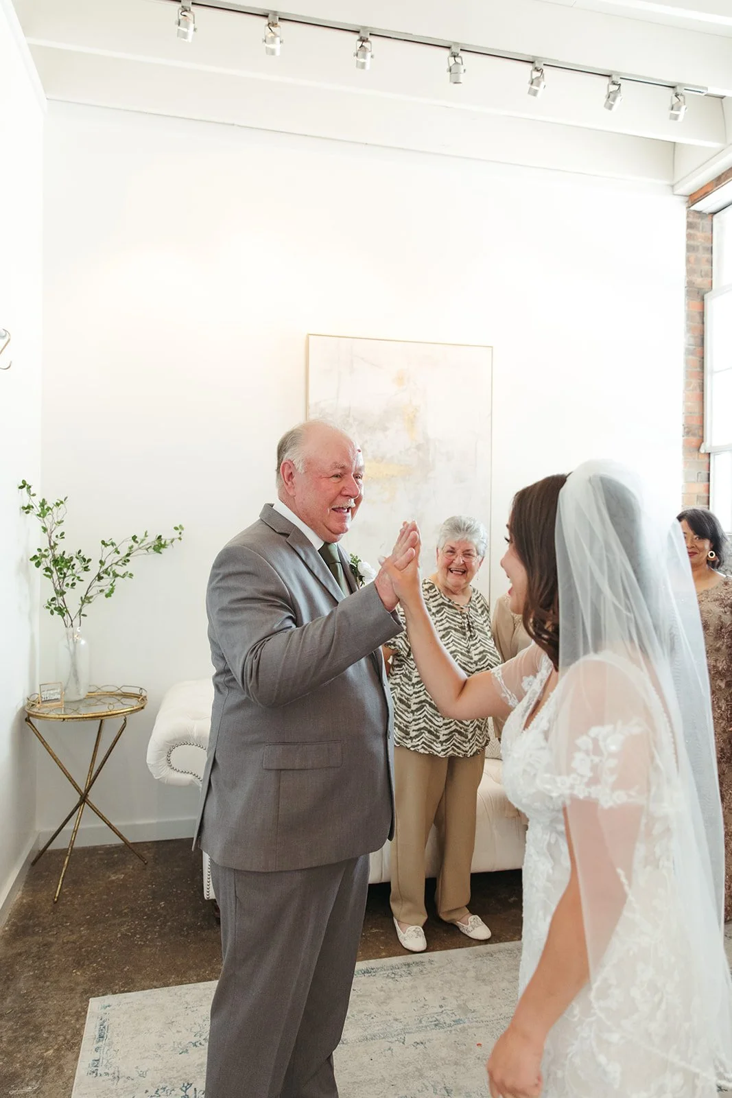 A bride and an older man, likely her father, greeting each other with a high-five during a wedding. Two women are smiling in the background in a bright, modern room.
