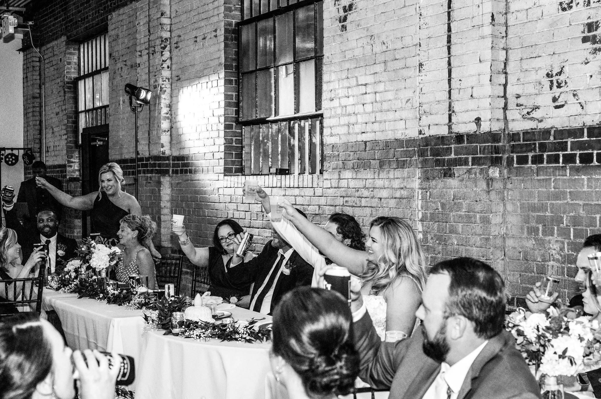 People celebrating at a wedding reception, raising glasses in a toast, seated at a decorated table against a brick wall background in black and white.