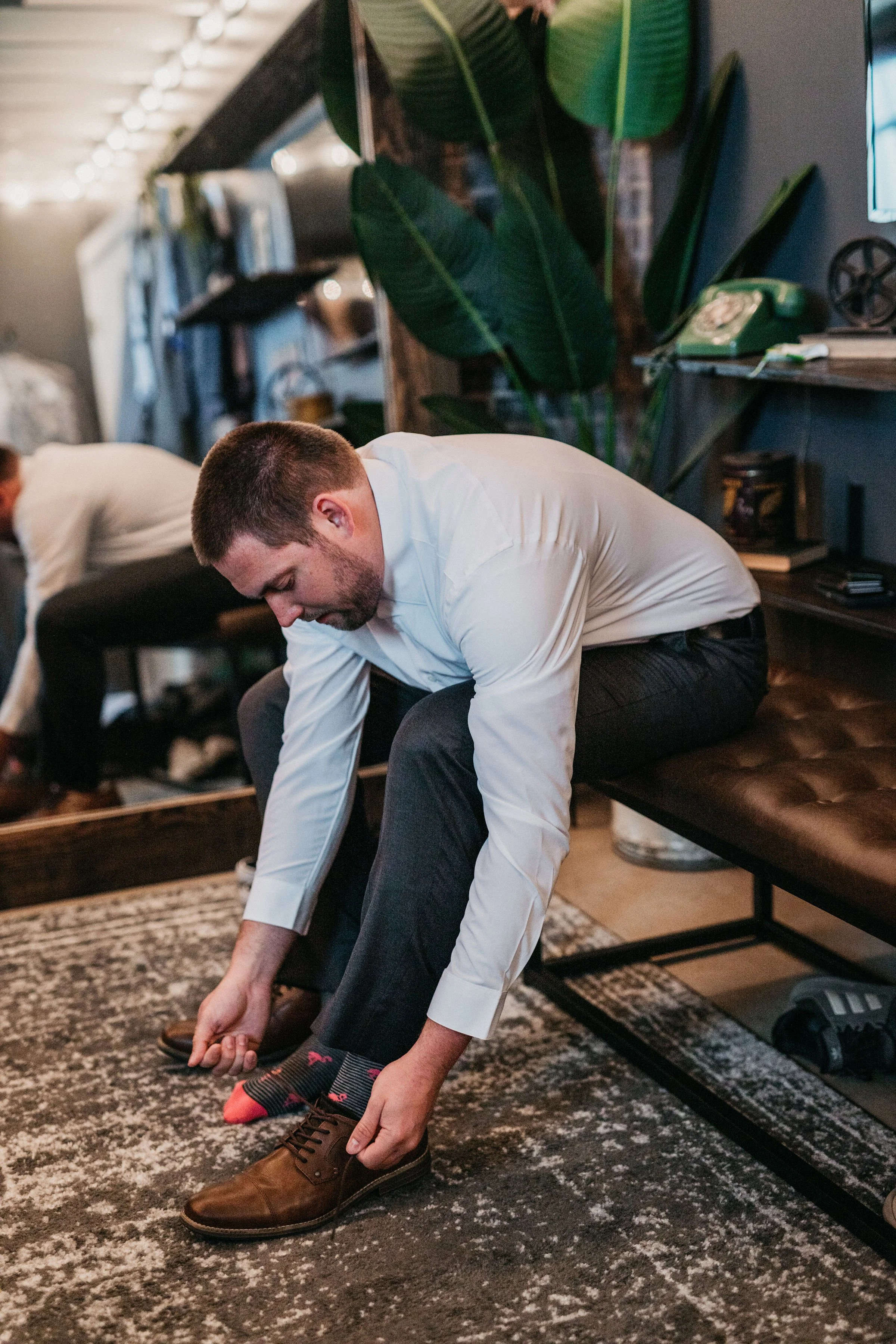 Man in white shirt and dark pants sitting on a bench tying his brown dress shoe in an indoor space with plants, books, and vintage decor.