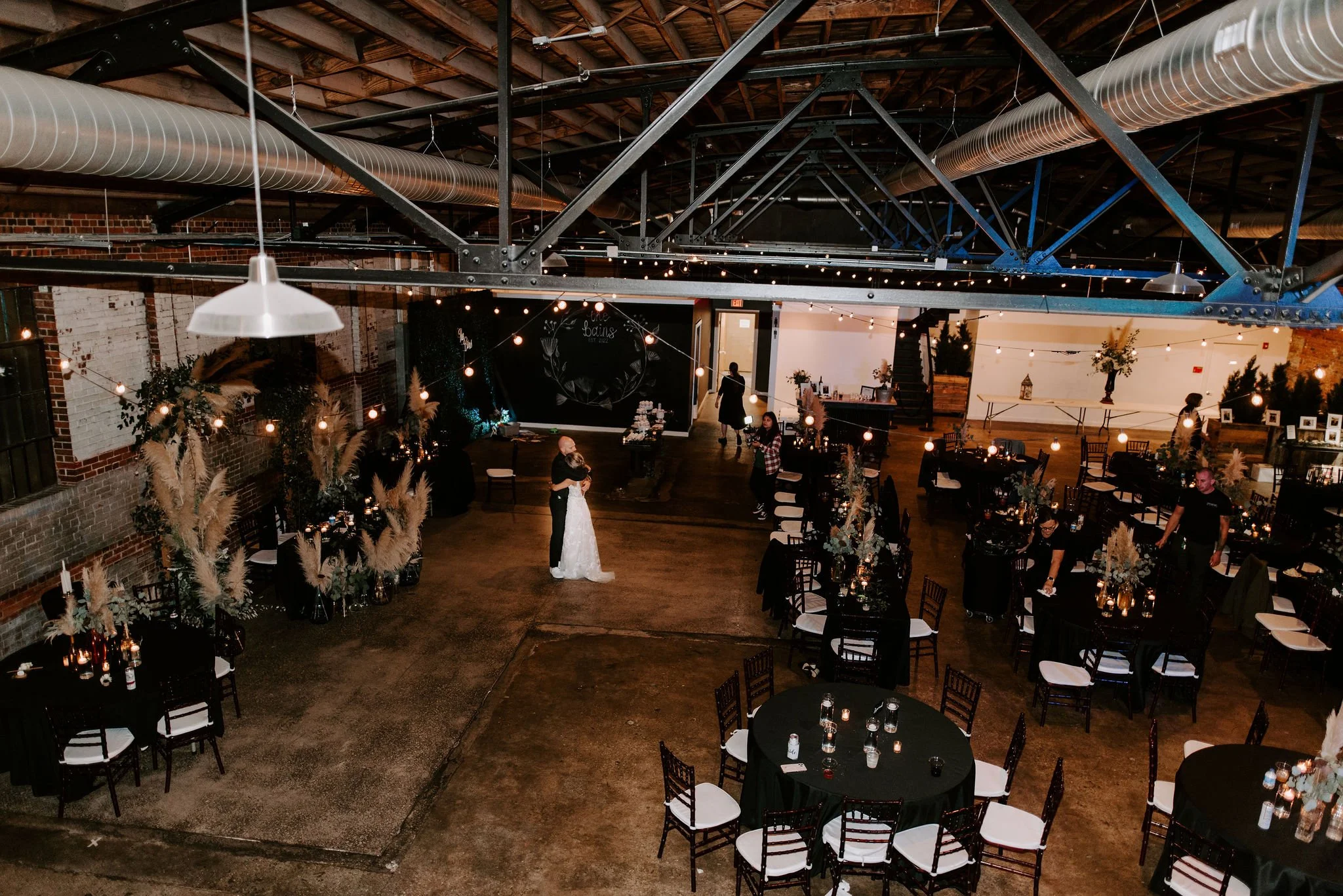 Wedding reception in industrial-style venue with string lights, decorated tables, and a bride and groom dancing.