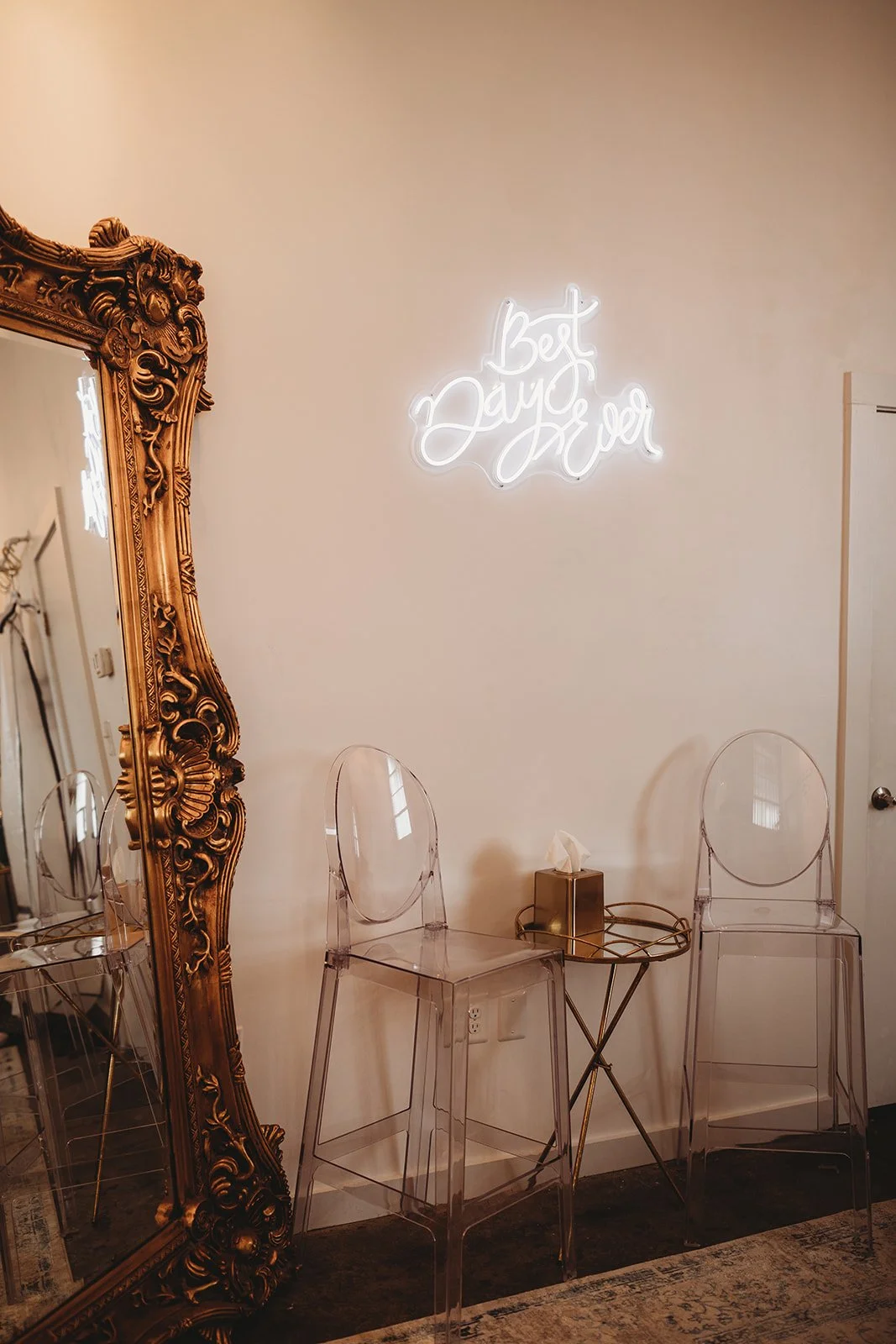 Decorative wall with lit neon sign saying 'Best Day Ever', vintage ornate mirror, and three clear acrylic chairs with a small gold side table holding a tissue box.