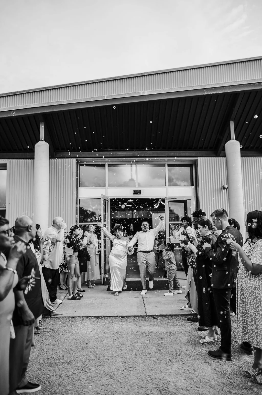 A black-and-white photo of a wedding celebration outside a building, with two people in wedding dresses holding hands and smiling as they exit, surrounded by guests cheering and taking photos, with confetti in the air.
