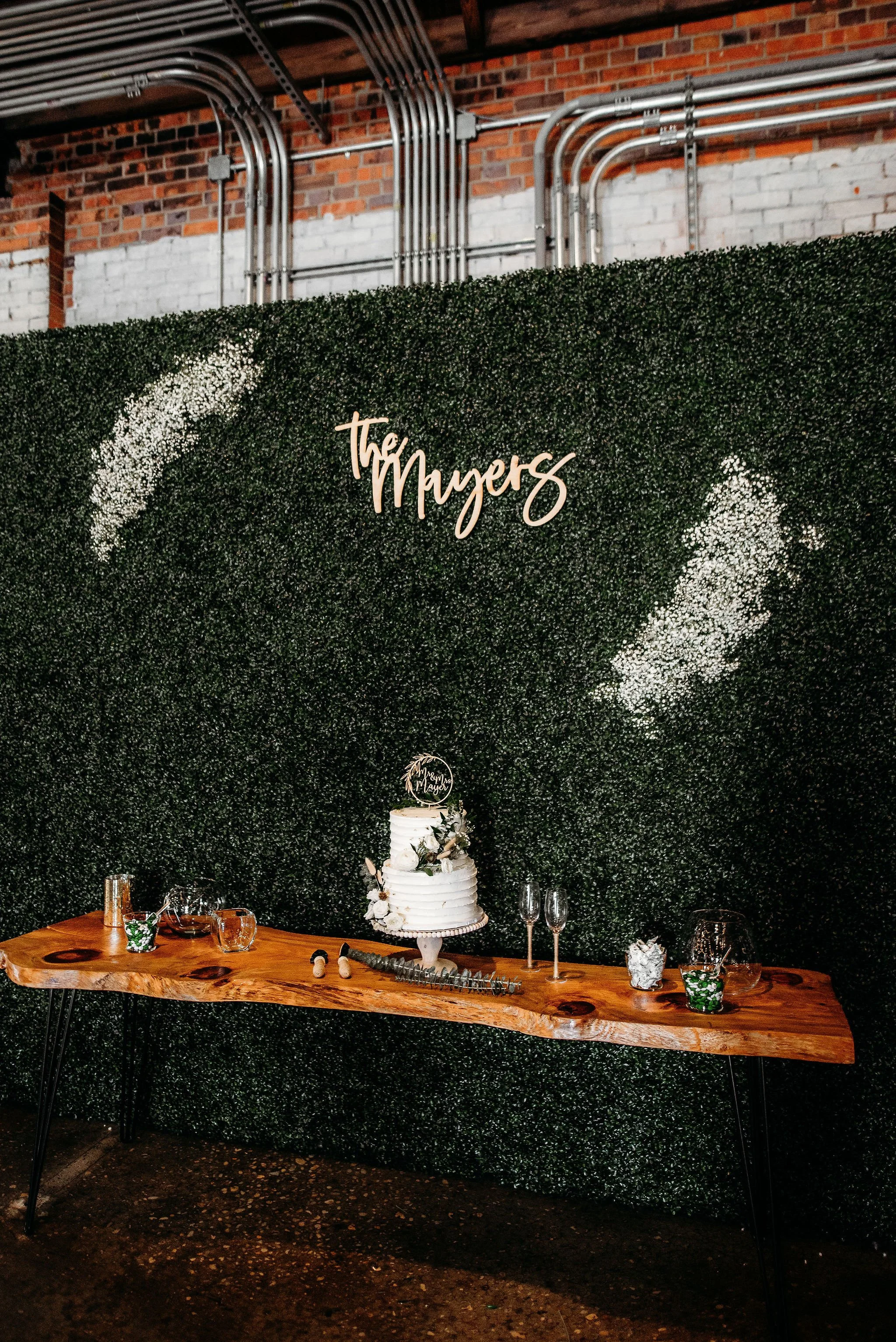 Wedding cake on a rustic wooden table with champagne flutes, glasses of water, and decorations, in front of a green hedge wall with white text that reads 'The Myers'.